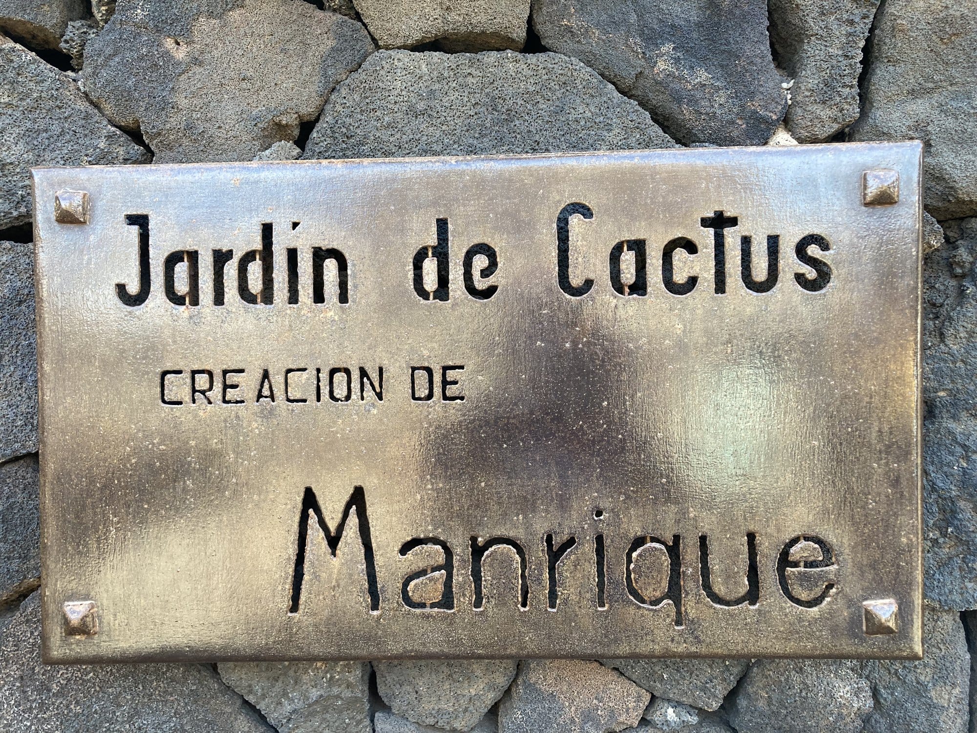 The brass entrance plaque of the Jardín de Cactus — "Creación de Manrique" — set into the black volcanic stone wall — Lanzarote
