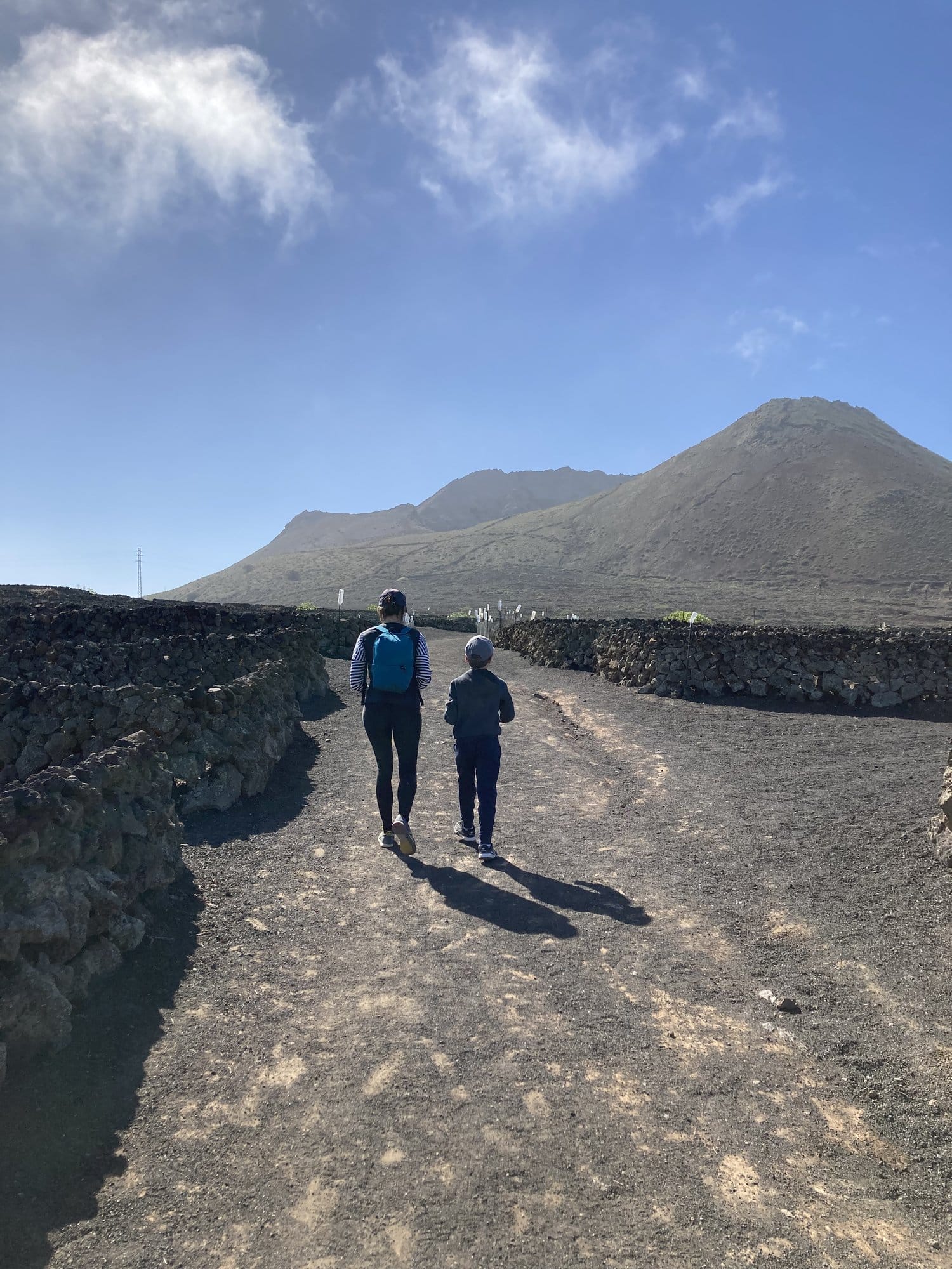 An adult and child walking along a volcanic stone path towards a mountain — lava stone walls lining the trail, clear blue sky — Lanzarote