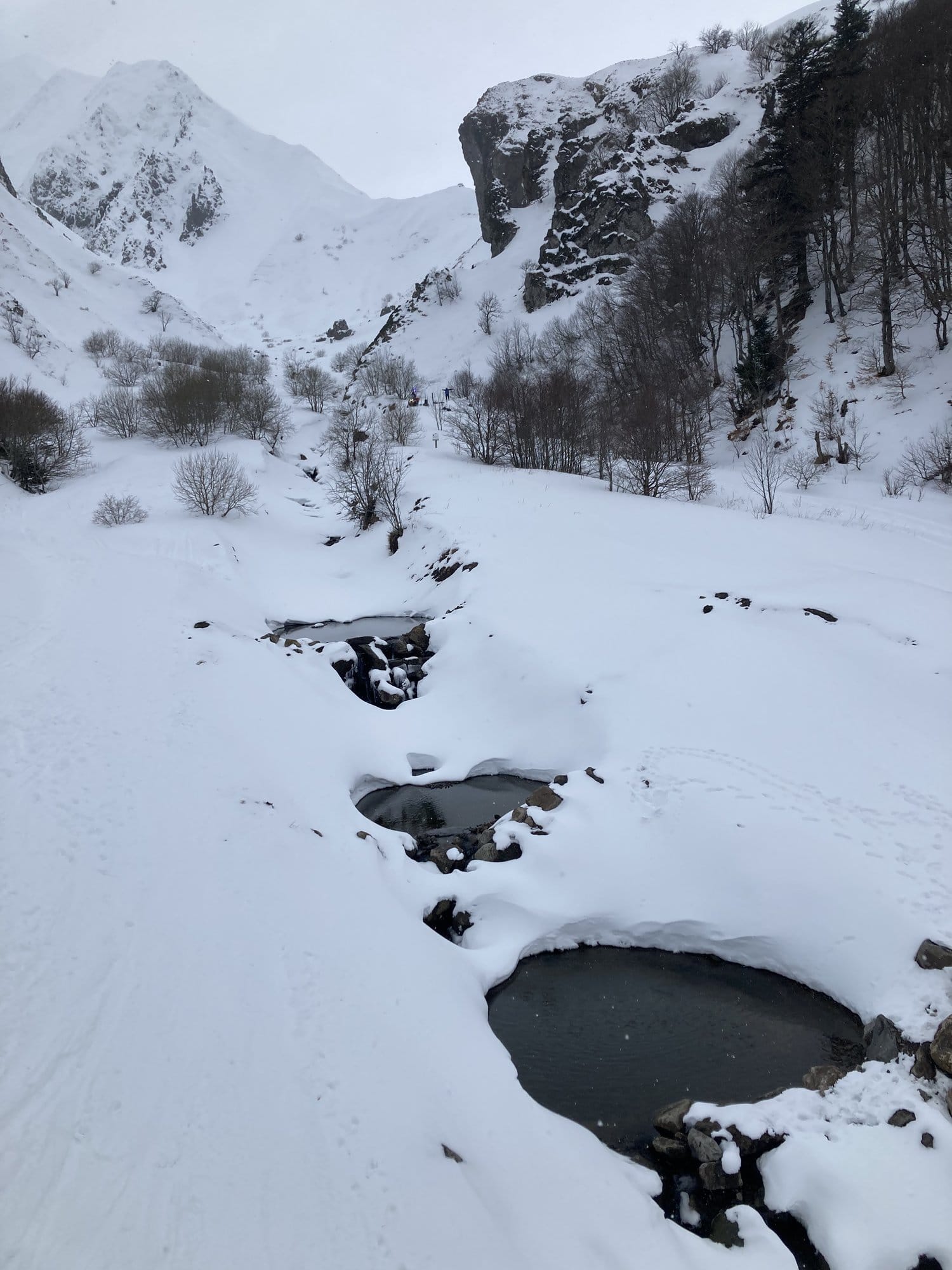 A frozen stream winding through the snow-covered Vallée de Chaudefour, with bare trees and rocky cliffs rising in the background — Auvergne, France
