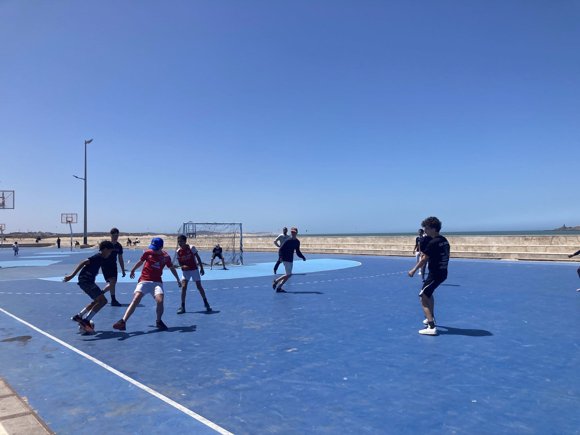 Kids playing a match on the blue port-side football pitch with the old ramparts and a painted mural behind — Essaouira, Morocco