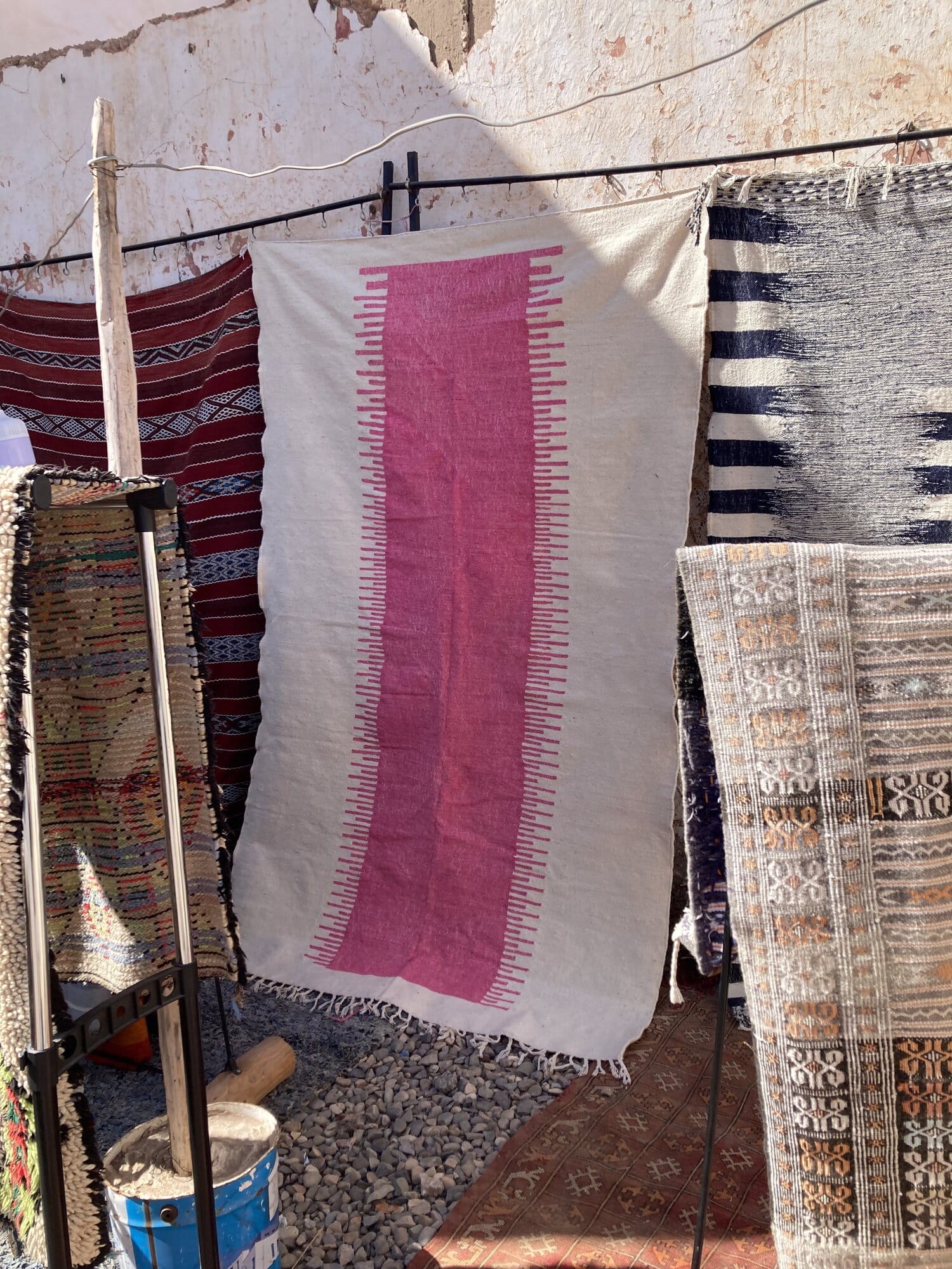 A row of suspended Berber kilim rugs in a Mirleft stall — a cream-and-fuchsia centrepiece with black-and-white and red kilims alongside — Mirleft, Morocco