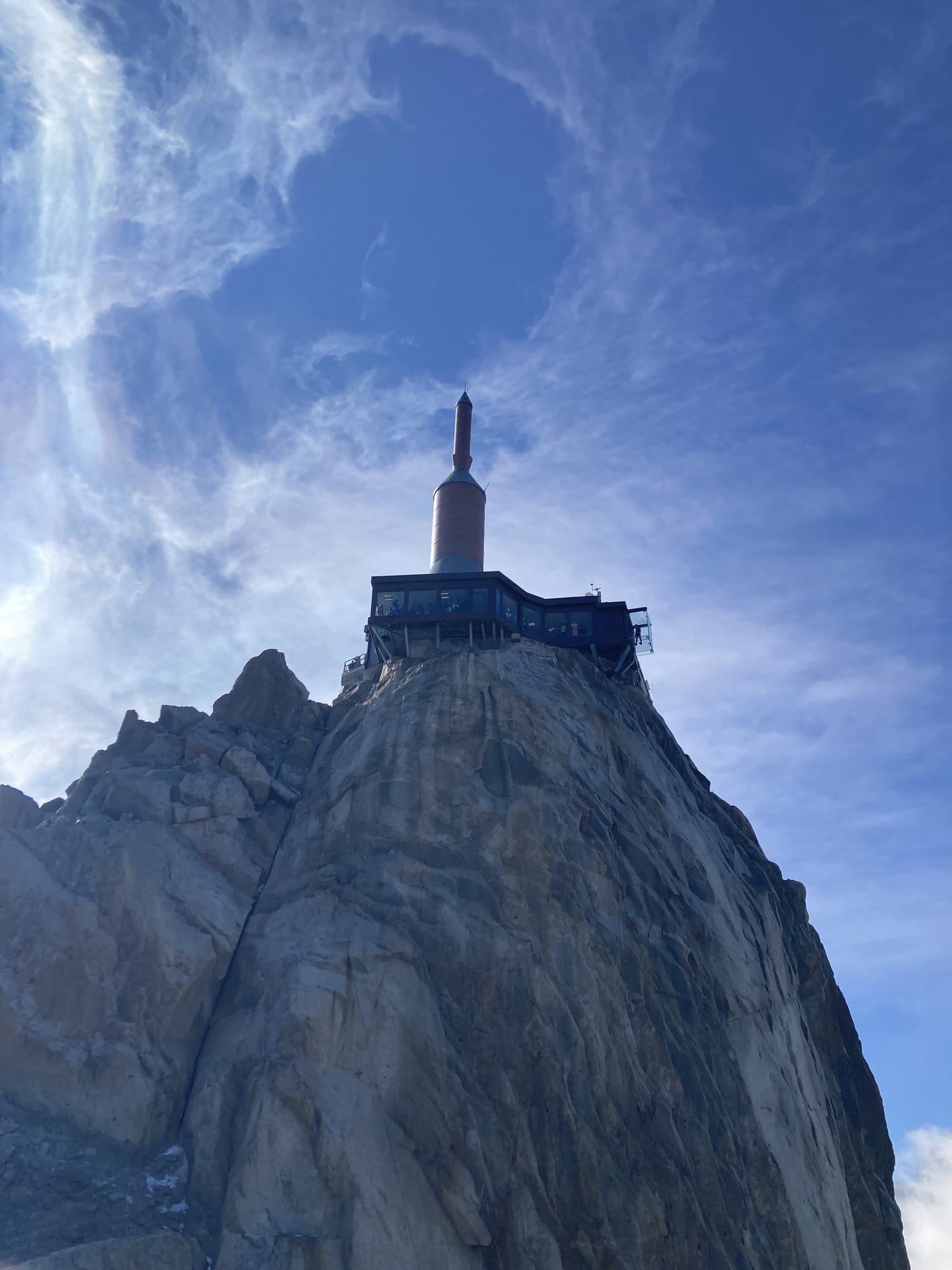 The iconic summit spire of the Aiguille du Midi viewed from below, with its antenna and observation platform perched on a granite needle against dramatic clouds — Chamonix-Mont-Blanc, France