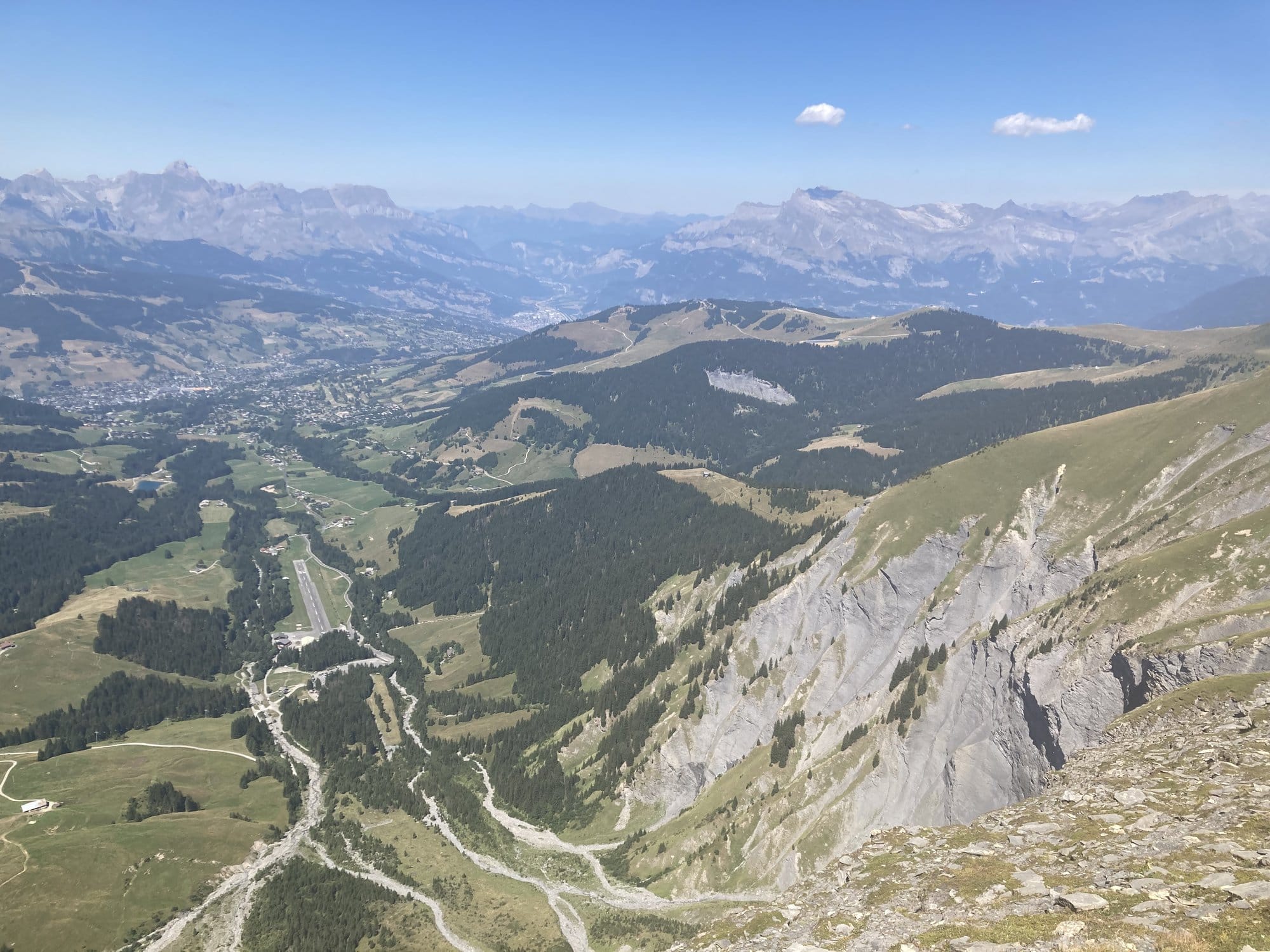 View from a high summit at 2,418m looking down over the valley around Megève, with rocky cliffs in the foreground and the Aravis range on the horizon — Megève, France
