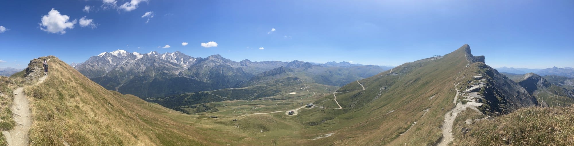 Ultra-wide panoramic view from a ridgeline col at 2,384m showing Mont-Blanc, green alpine grasslands, and hiking trails — above Megève, France