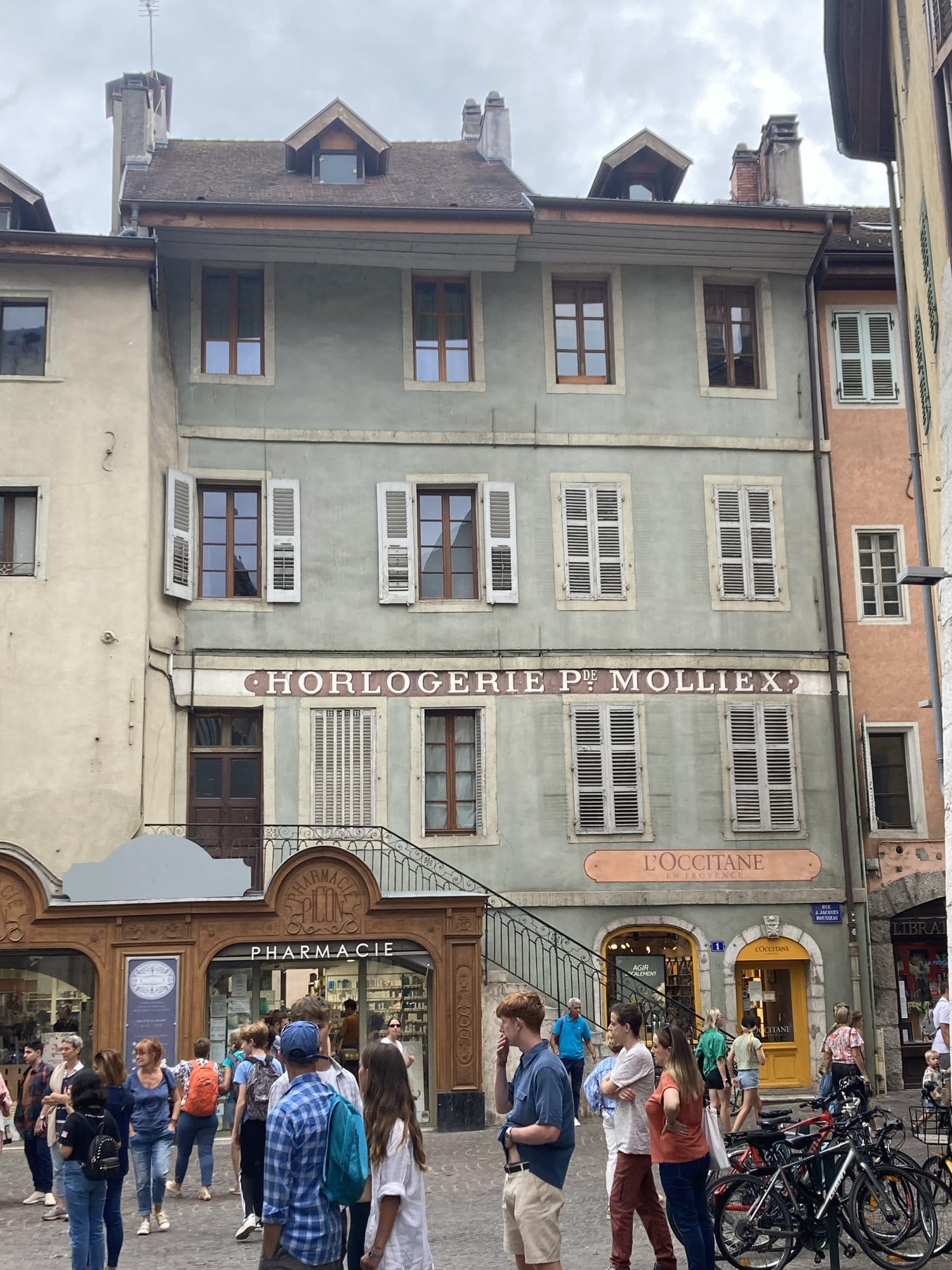 Historic building in Annecy's old town with the original painted signage of Horlogerie Molliex, alongside an L'Occitane boutique and a pharmacy — Annecy, France