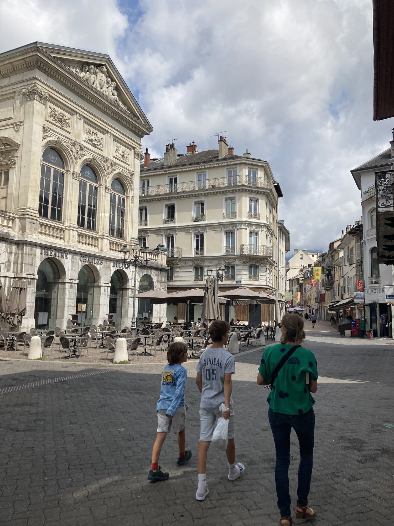A family walking through the Place du Théâtre in Chambéry, with the classical theatre building and café terraces visible — Chambéry, France