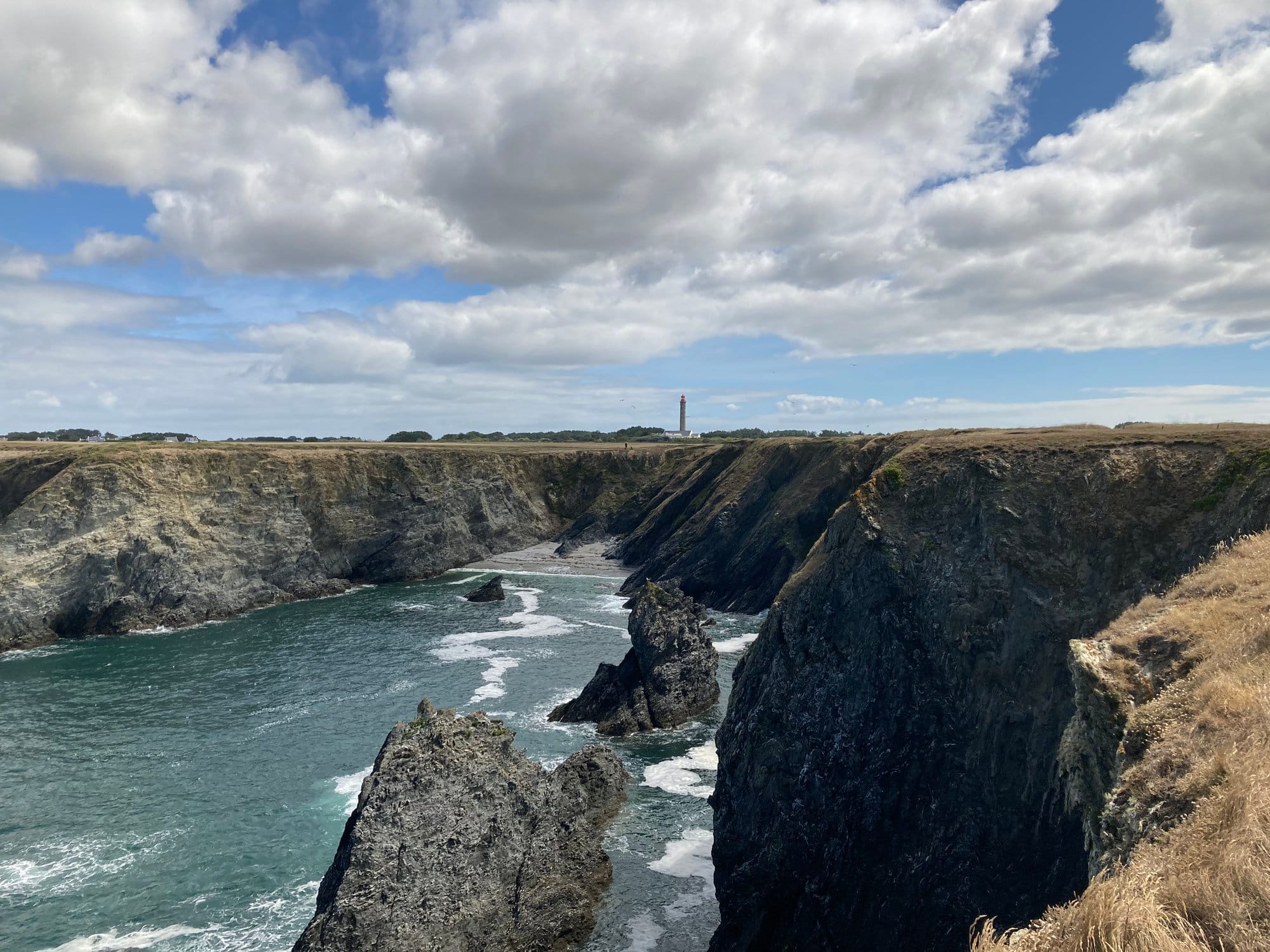 Rugged coastal cliffs with deep inlets cut into the rock, the Grand Phare lighthouse visible on the distant headland