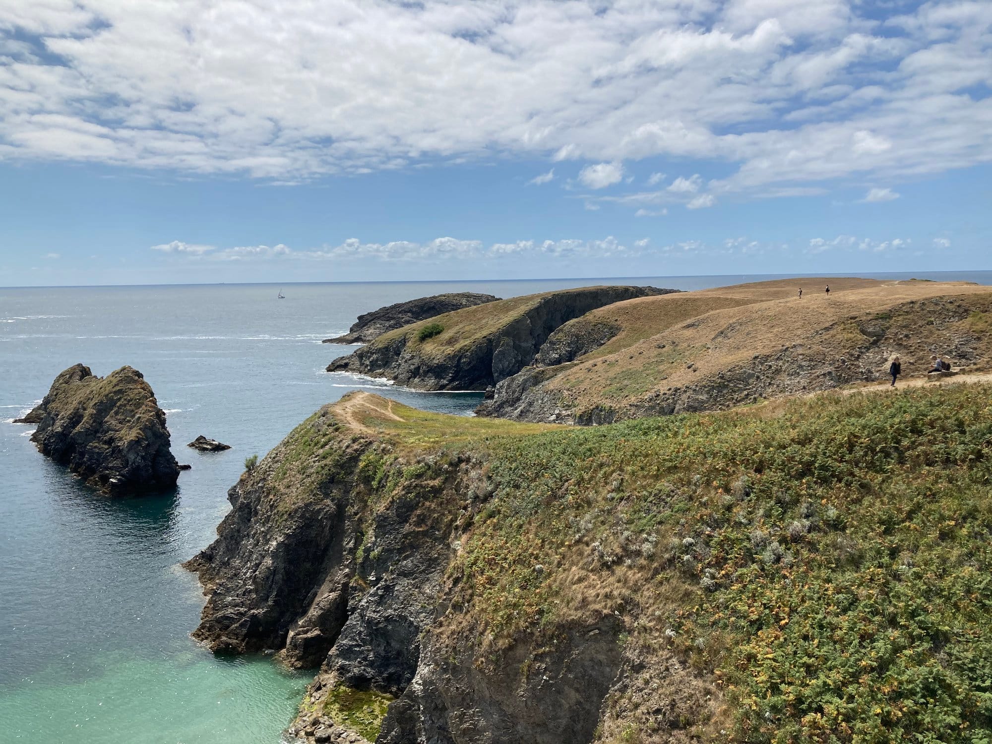 Cliff-top path with a wide view of a sheltered bay and headland, two small figures walking the trail, green scrub dropping to clear water