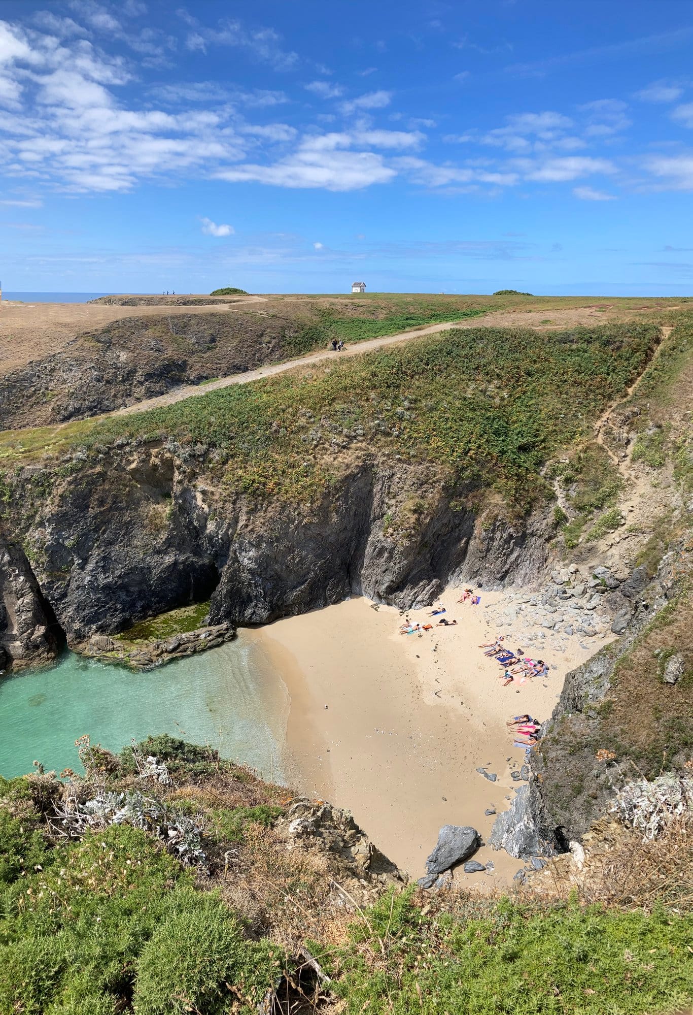 A hidden sandy beach in a clifftop bowl, a few sunbathers on the sand below, the Grand Phare lighthouse silhouetted on the headland