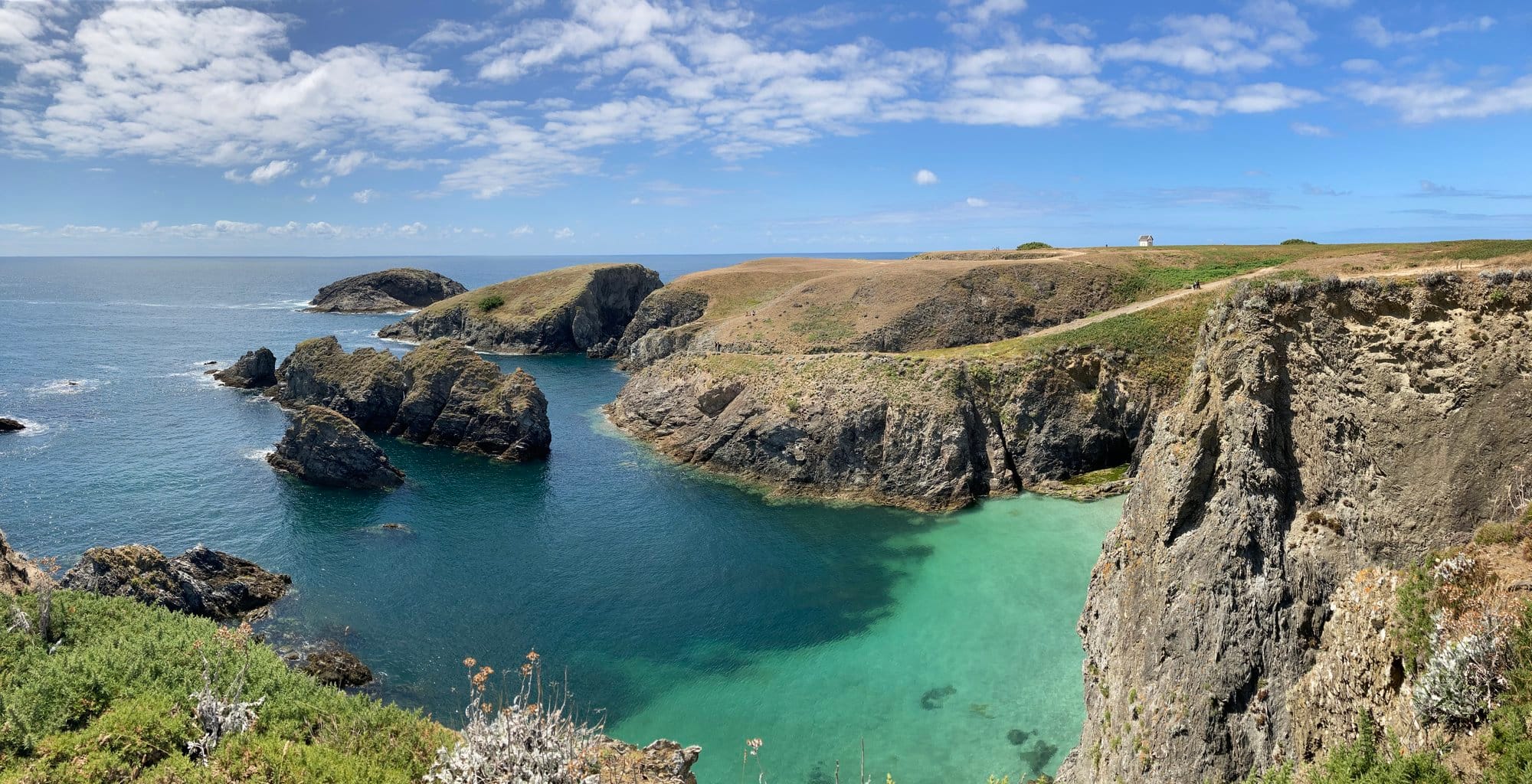 Wide panoramic view of a turquoise cove with jagged rocky headlands on either side, calm clear water catching the afternoon sun
