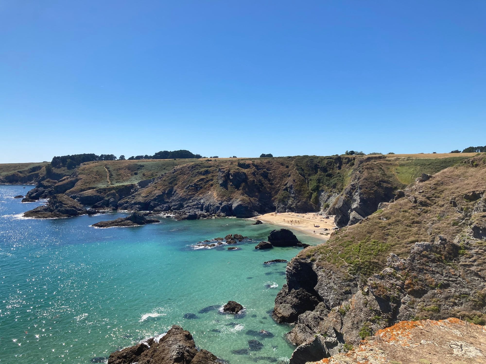 A wild cove with bright turquoise water, rocky outcrops framing a small secluded sandy beach, cliffs rising on either side — one of the island's hidden gems