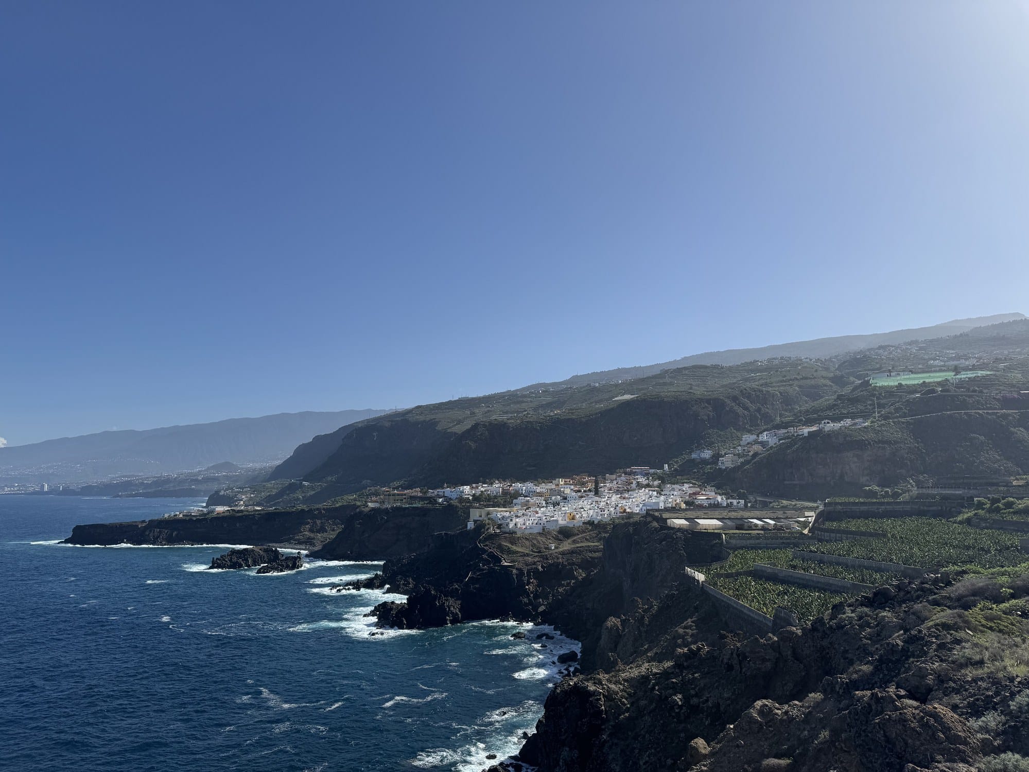 Vista panoramica da un mirador su un villaggio bianco arroccato sopra scogliere basaltiche, con piantagioni di banane che scendono verso un'insenatura turchese dell'Atlantico — San Marcos, nord di Tenerife