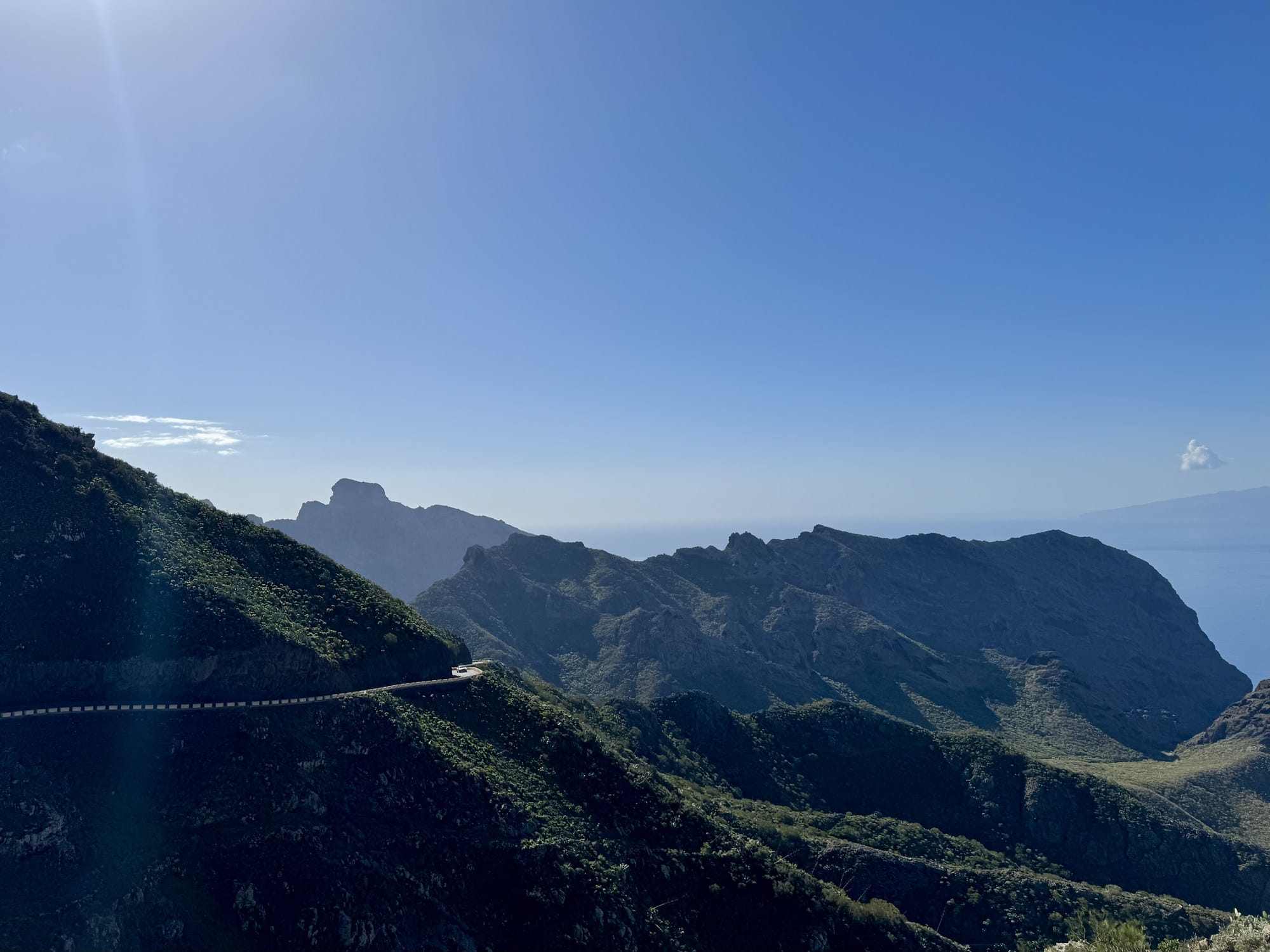 A winding mountain road cut into the dry, sharp ridges of the Teno massif, with La Gomera visible on the horizon — Tenerife