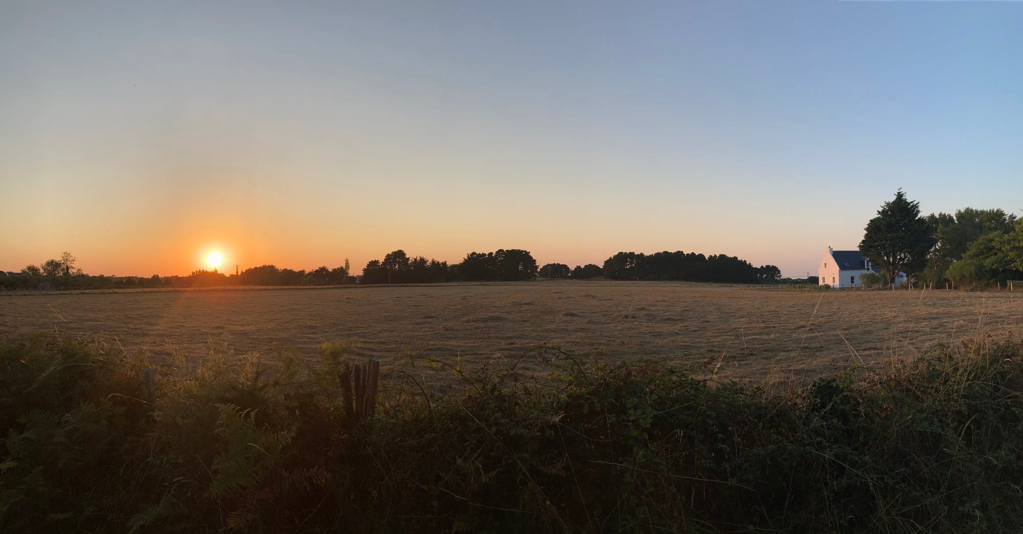 Golden sunset over a wide Breton field, the silhouette of a whitewashed farmhouse to the right, trees glowing amber against an orange sky