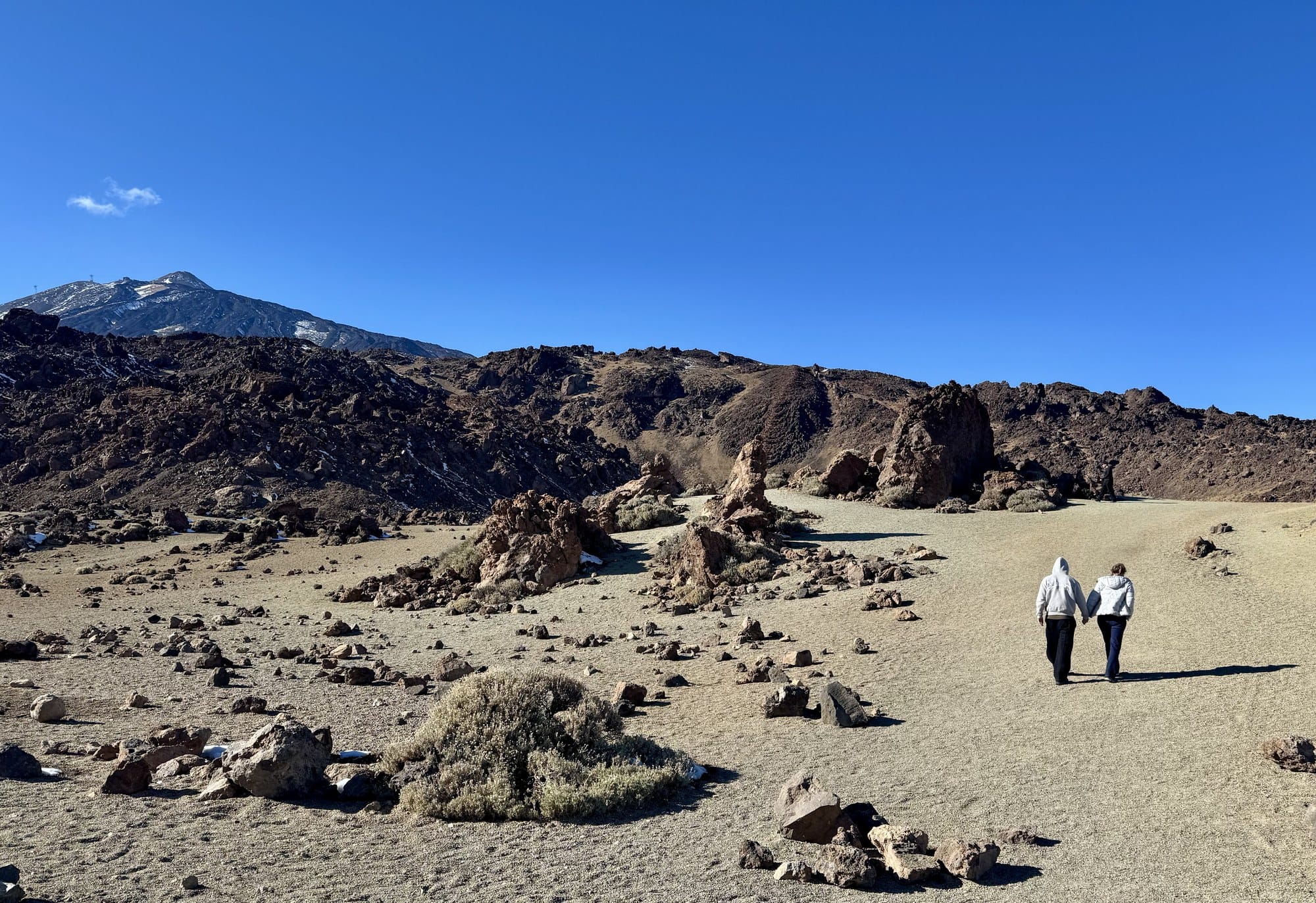 Two people walking hand in hand on the pale volcanic sand of the Minas de San José, with Teide rising behind — Teide National Park, Tenerife