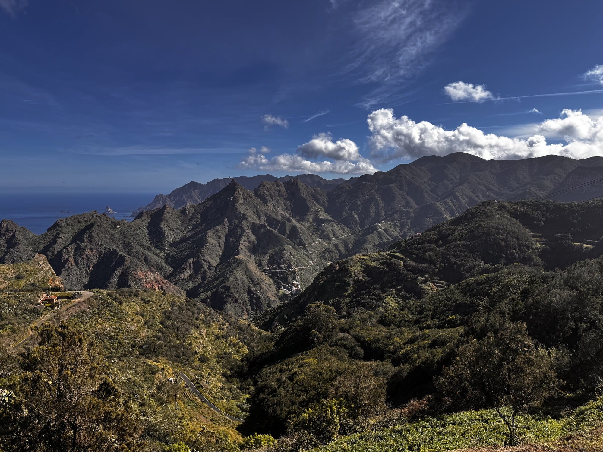 A panoramic view from an Anaga mirador: a jagged green ridge falling toward the Atlantic, with three small rocky islets on the horizon — Tenerife