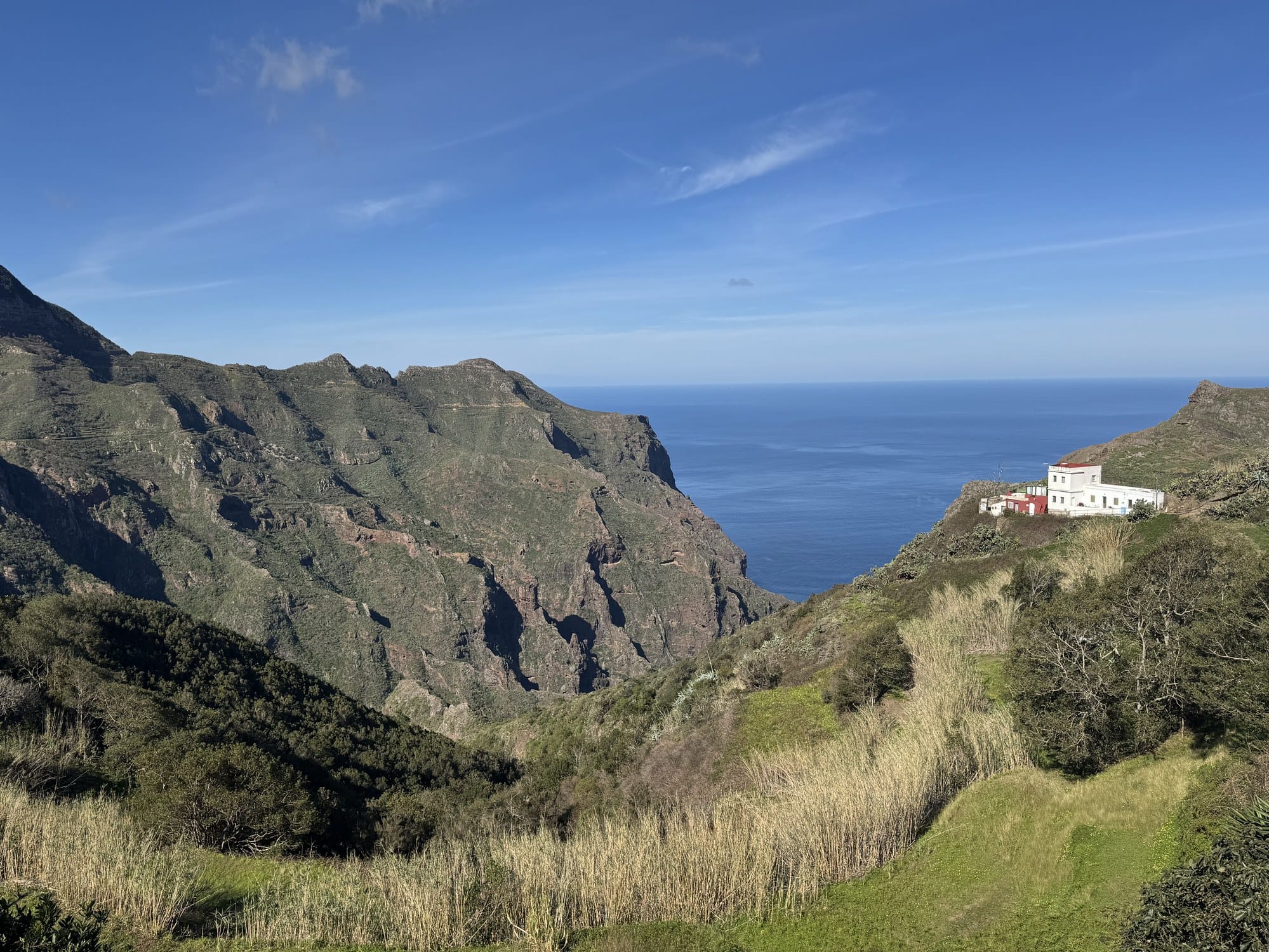 A single white house perched on a ridge above a deep green valley in Anaga, with the Atlantic Ocean in the background — Tenerife
