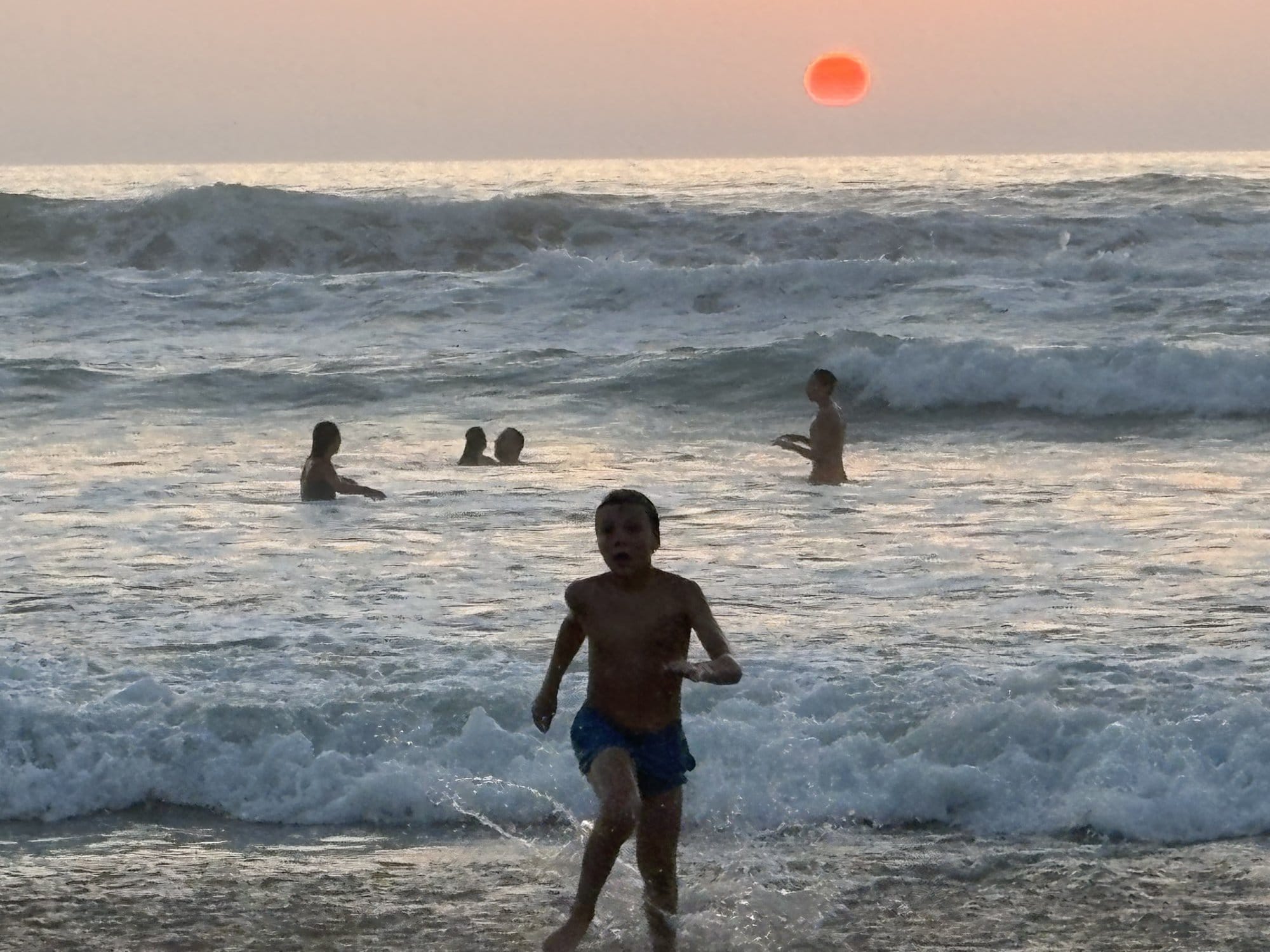 A child running through the waves at sunset on Bidart beach, with the sun glowing red-orange above the horizon — French Basque Country