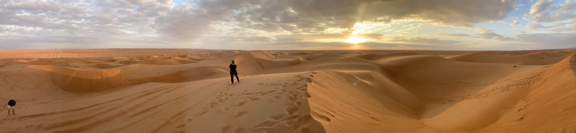 Panoramic sunset over the vast sand dunes with a lone figure silhouetted on the crest โ Wahiba Sands, Oman