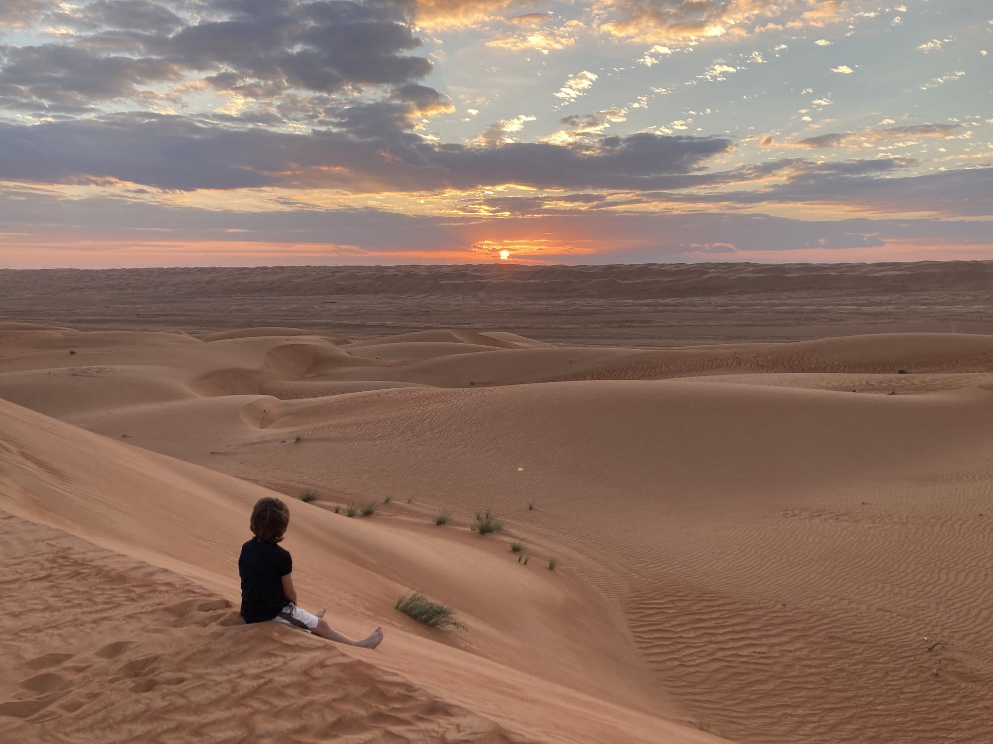 Child sitting on a sand dune watching the sunset, seen from behind โ Wahiba Sands, Oman