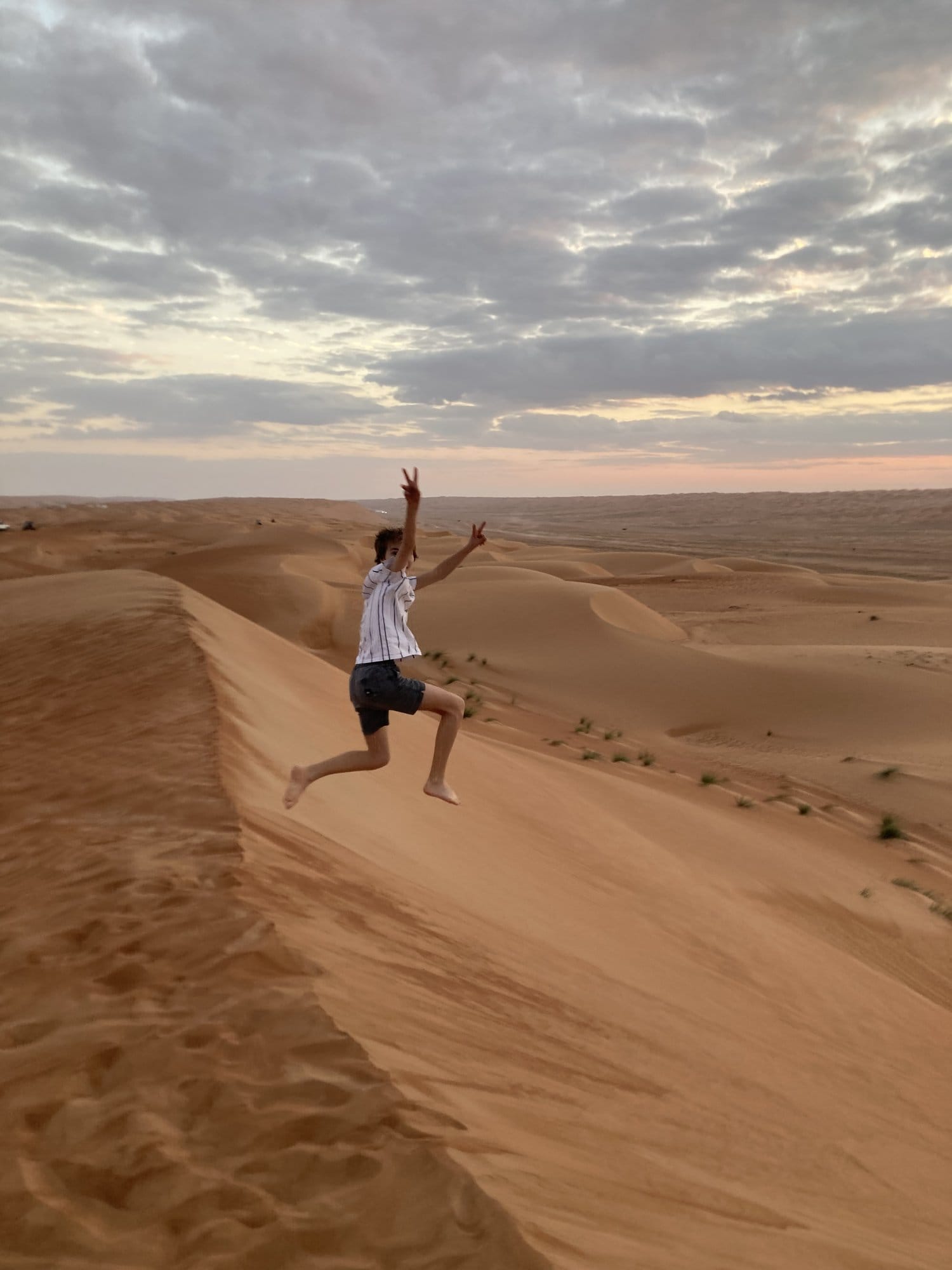 Child jumping with arms raised on the crest of a dune, silhouetted against the sunset โ Wahiba Sands, Oman