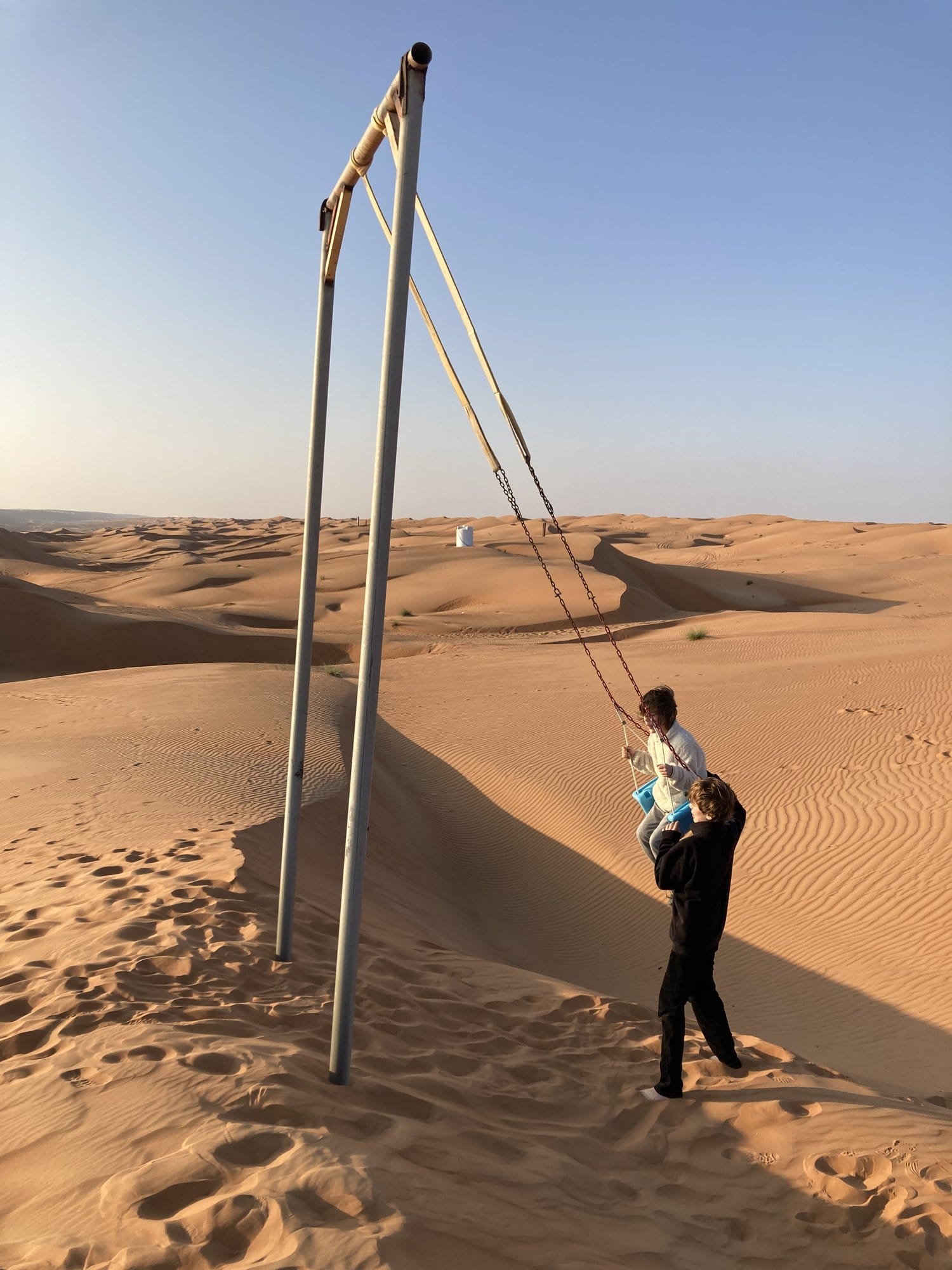 A swing set standing alone in the vast desert, with figures using it against the dune backdrop โ Wahiba Sands, Oman