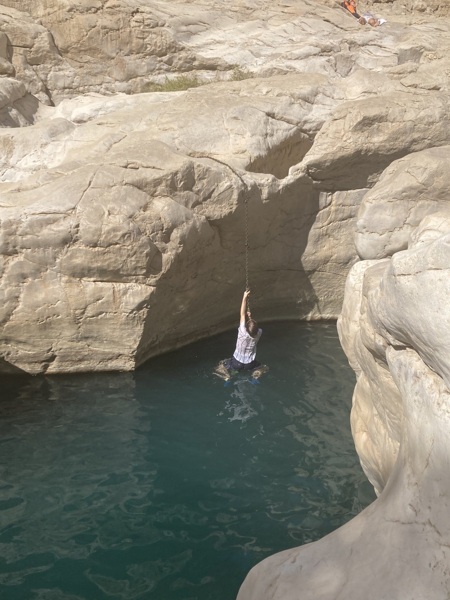 Person swinging on a rope over a turquoise wadi pool between rock walls — Wadi Bani Khalid, Oman