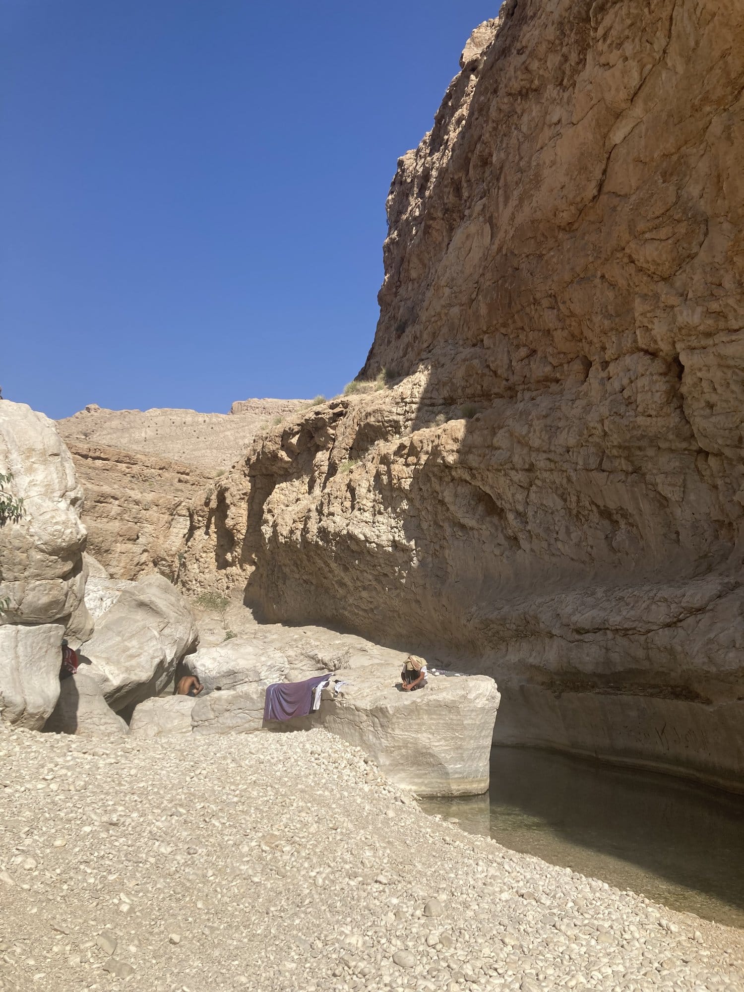 Wide pool at the base of tall ochre cliffs with people relaxing on the rocks — Wadi Bani Khalid, Oman