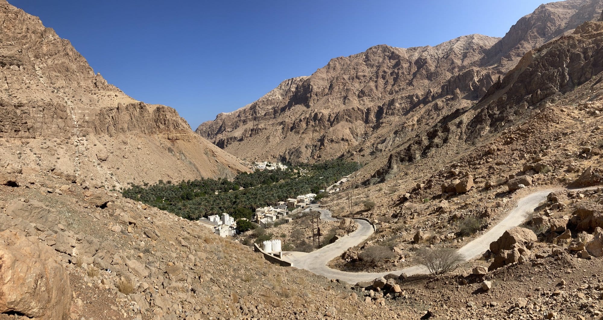 Panoramic view of the winding road descending into the Wadi Tiwi valley with its palm-filled village below — Sur, Oman