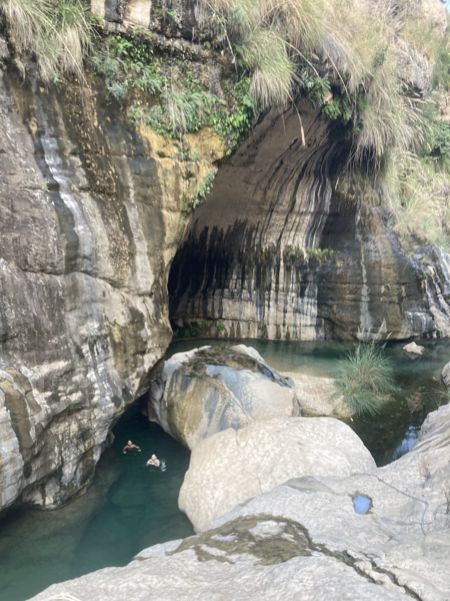 Grotto with curtain stalactites above a turquoise pool with swimmers below — Wadi Tiwi, Oman