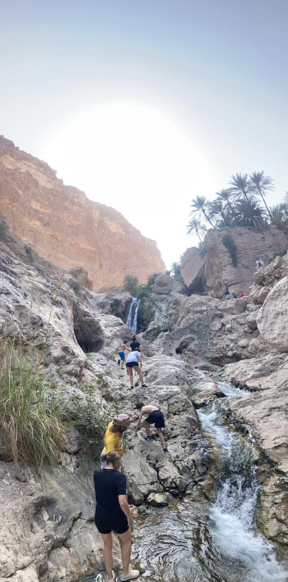 Family scrambling up rocks toward a waterfall, palm trees and canyon walls above — Wadi Tiwi, Oman