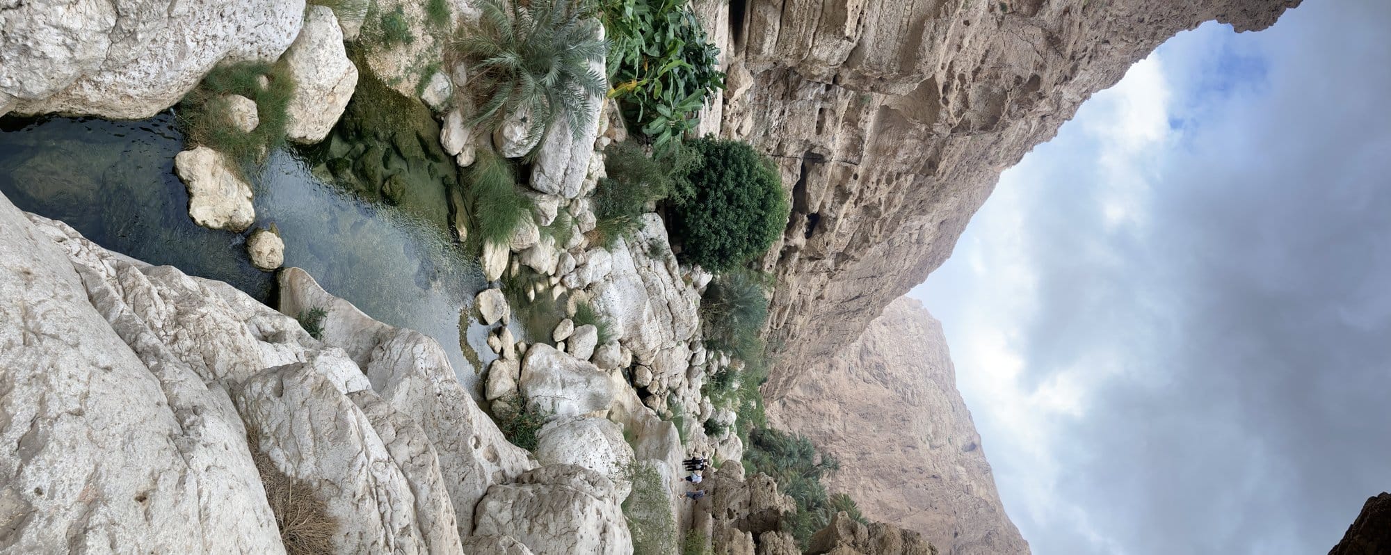 Panoramic view of a turquoise pool under a massive rock overhang with palms — Wadi Shab, Oman