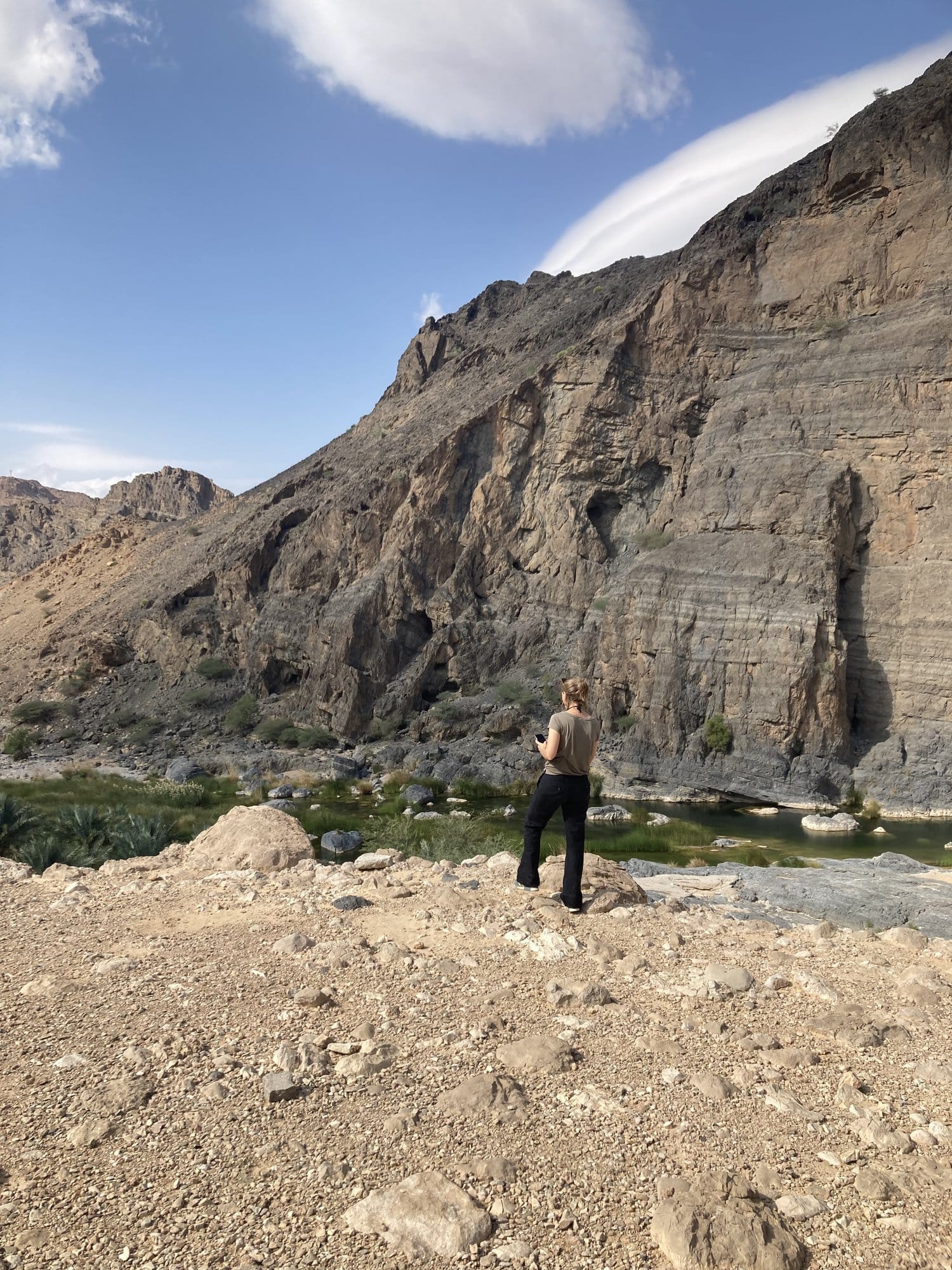Figure standing before a massive striated cliff face beside a green stream — Al Qābil, Oman