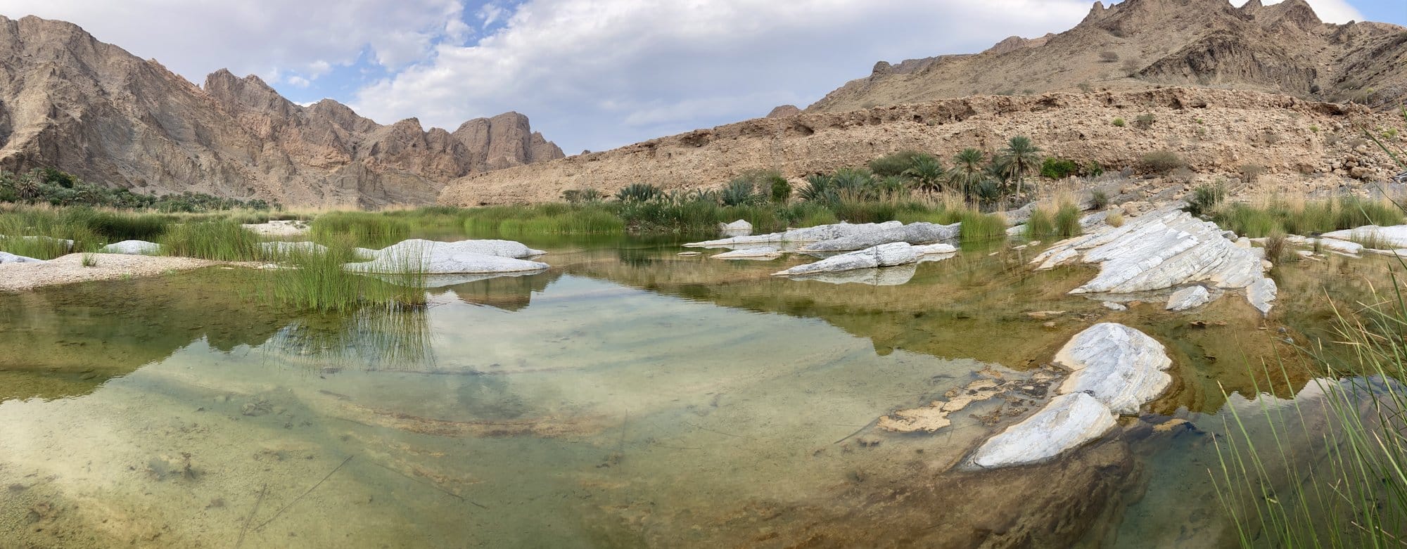 Panoramic pool with reeds, white rocks, and jagged mountains reflected in the water — Al Qābil, Oman