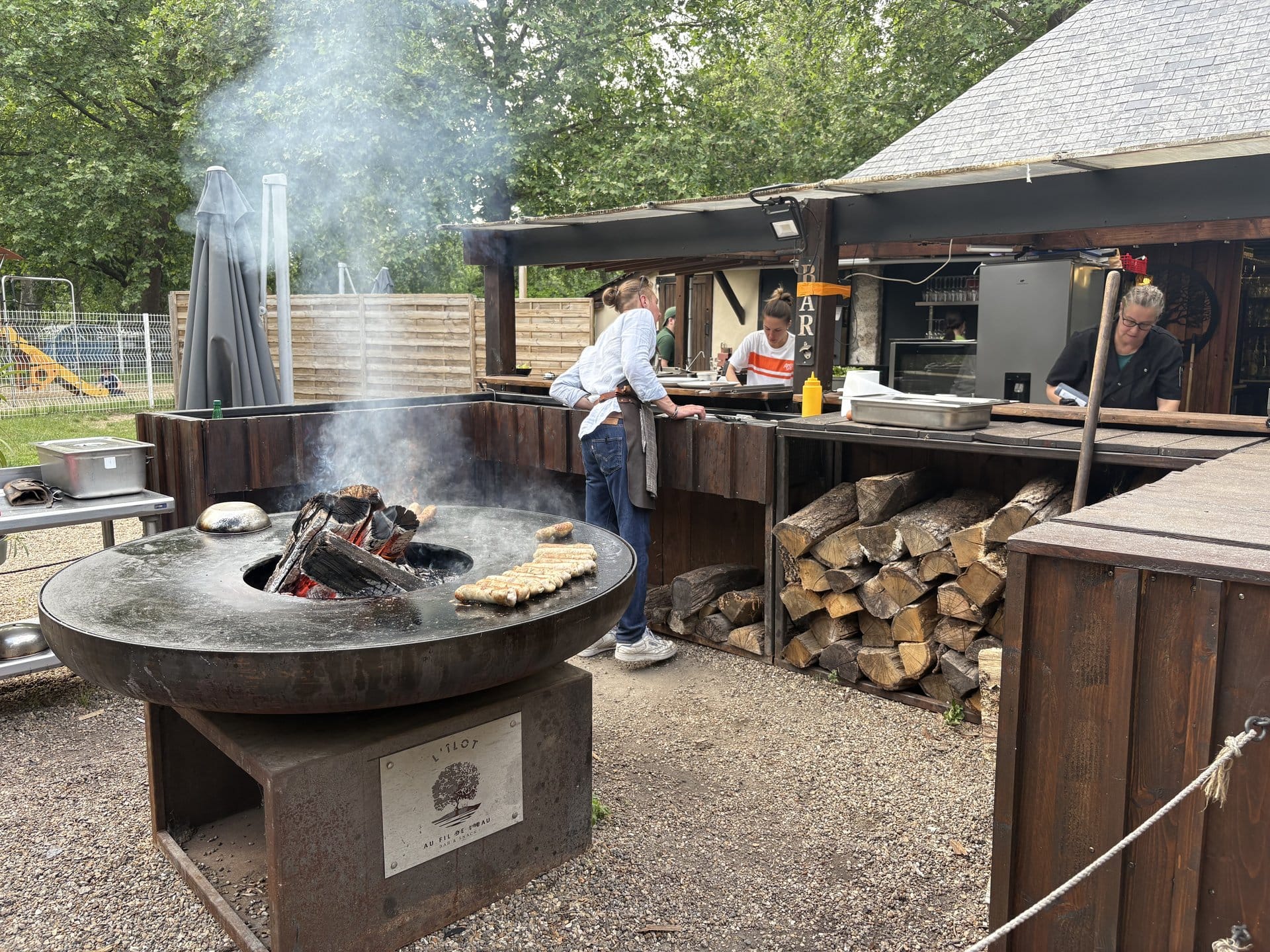 The round VIVON brasero at L'Îlot d'Azay cooking golden galettes around its rim, a chef in white shirt and blue apron flipping them, stacked logs behind