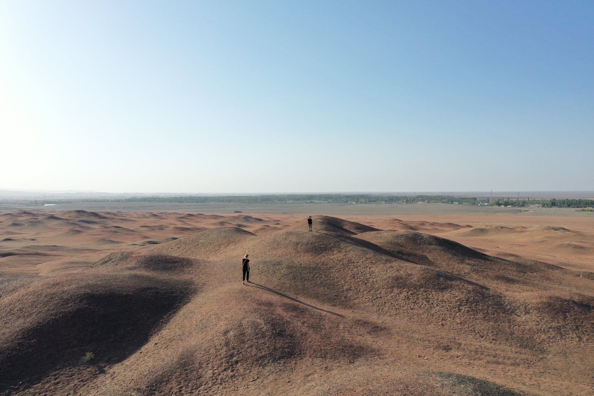 Rolling ochre desert hills with two tiny figures on a ridge and the green oasis on the horizon. Shot with a DJI Mavic 2 Pro.