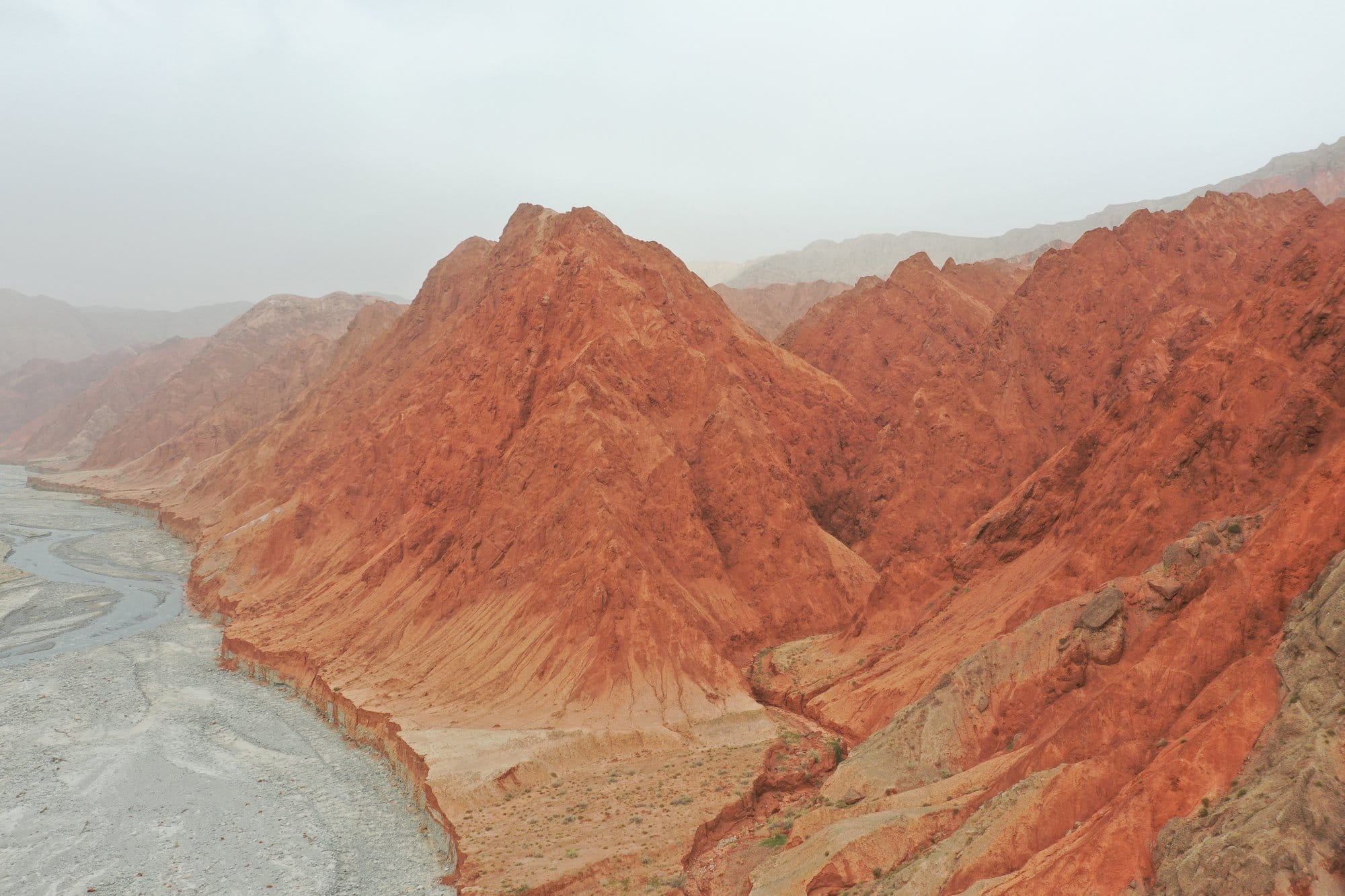 The red canyon at Ghez, orange ridges dropping to the pale braided riverbed in a haze of dust. Shot with a DJI Mavic 2 Pro.