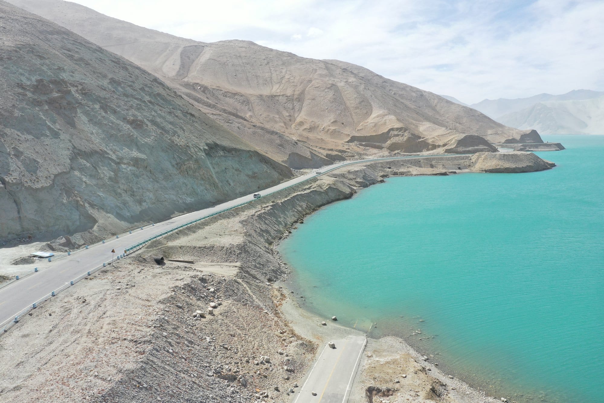 The mountain road winding along the edge of a turquoise lake between bare grey slopes. Shot with a DJI Mavic 2 Pro.