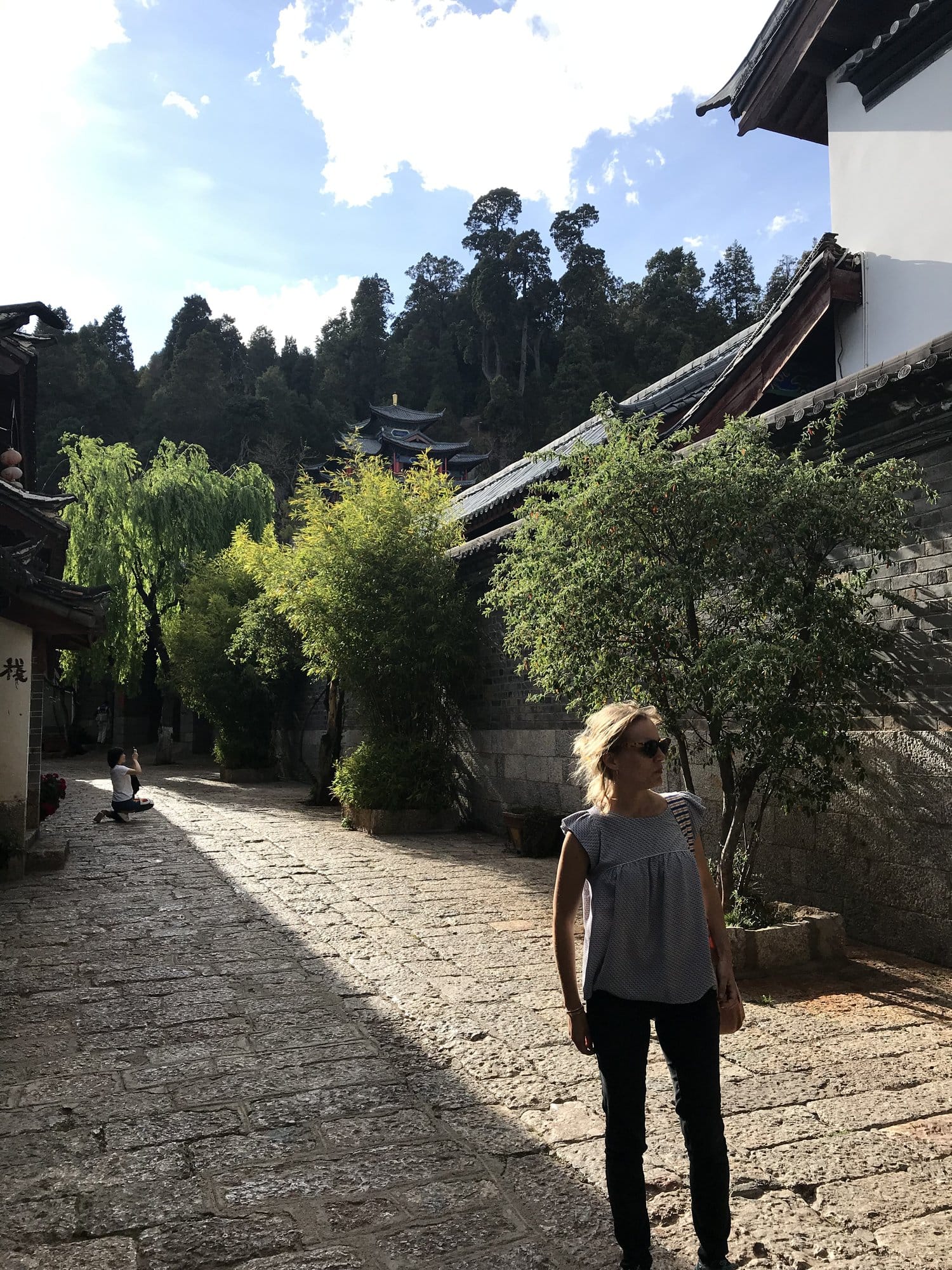 A quiet cobbled lane along a white wall, temple roof in the pines above.