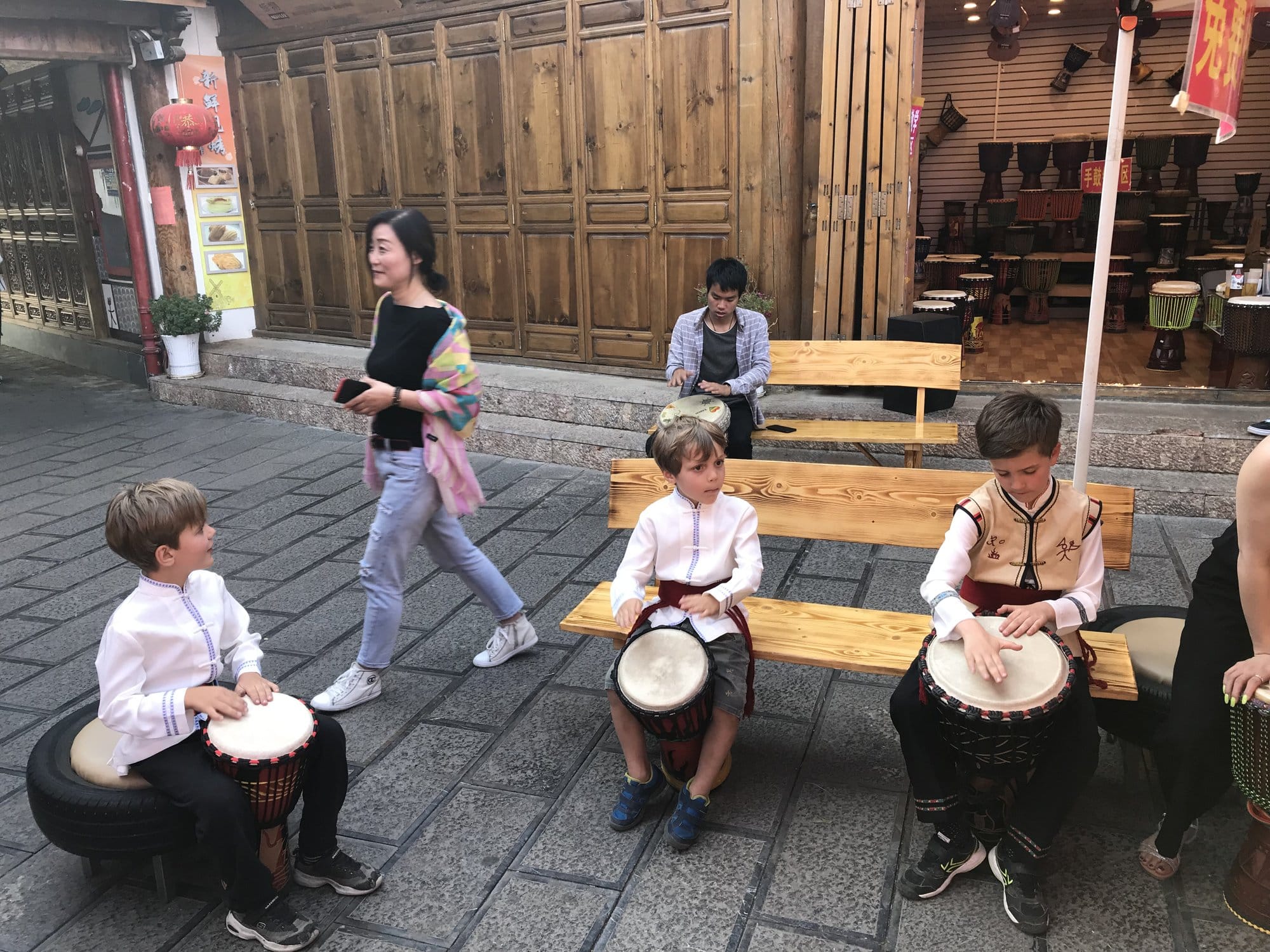 Drums set out on stools outside a workshop, children playing them in the street.