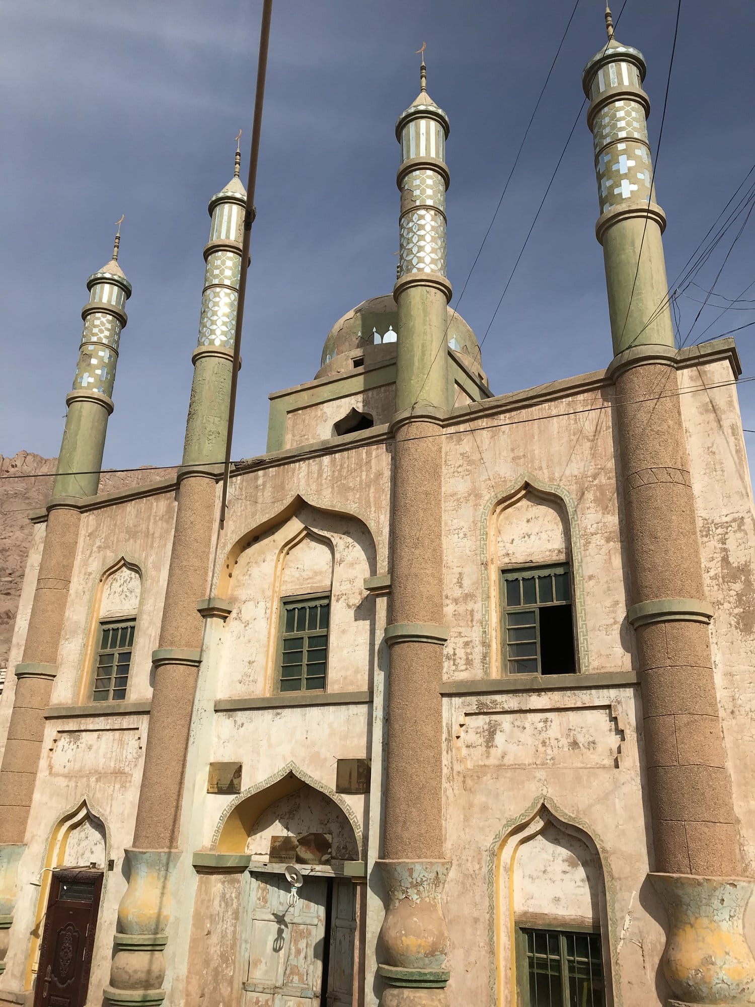 The village mosque from below: four green minarets with white patterning above worn brick and render.