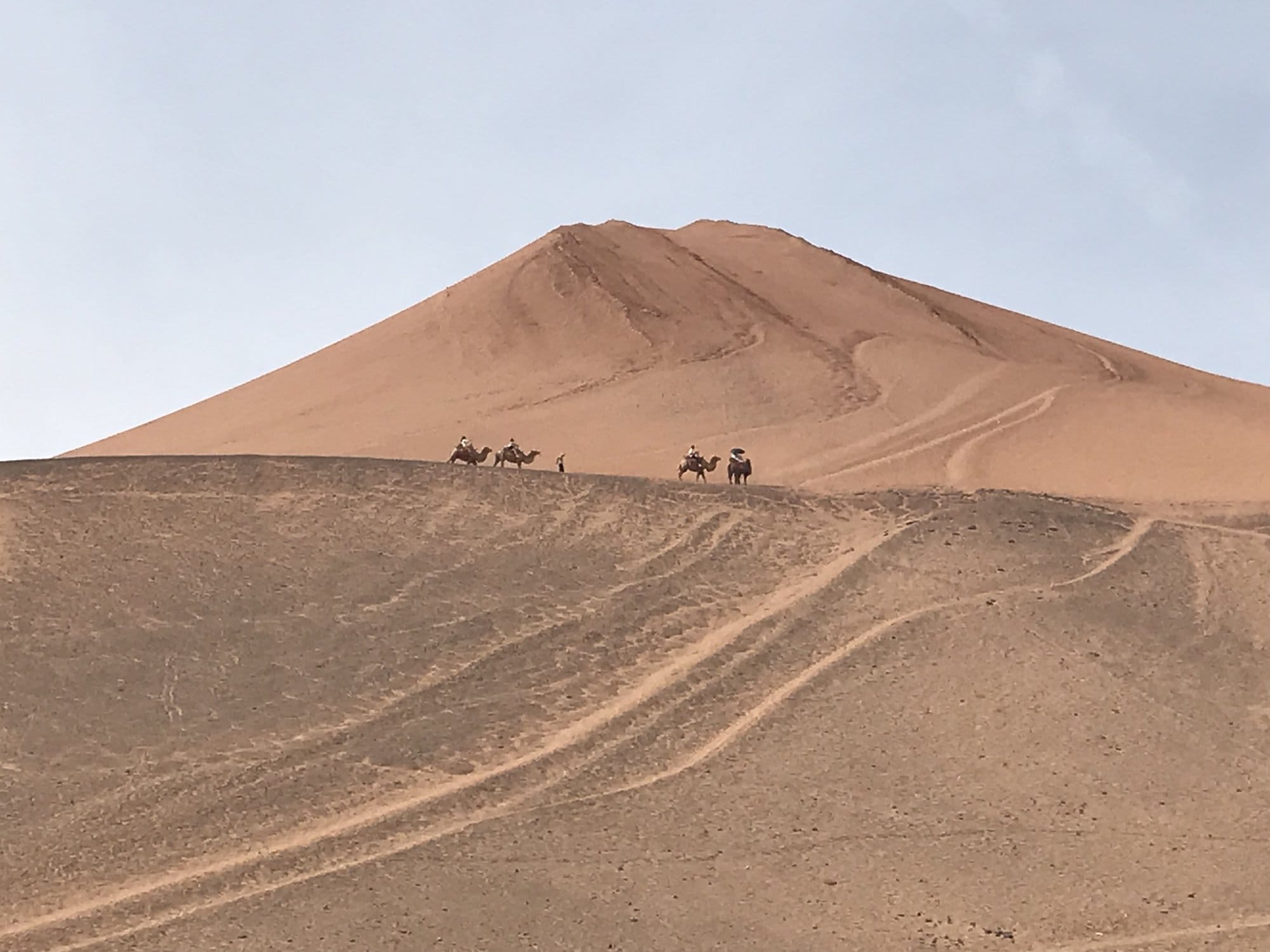 A conical dune with distant figures on camels moving along the crest.