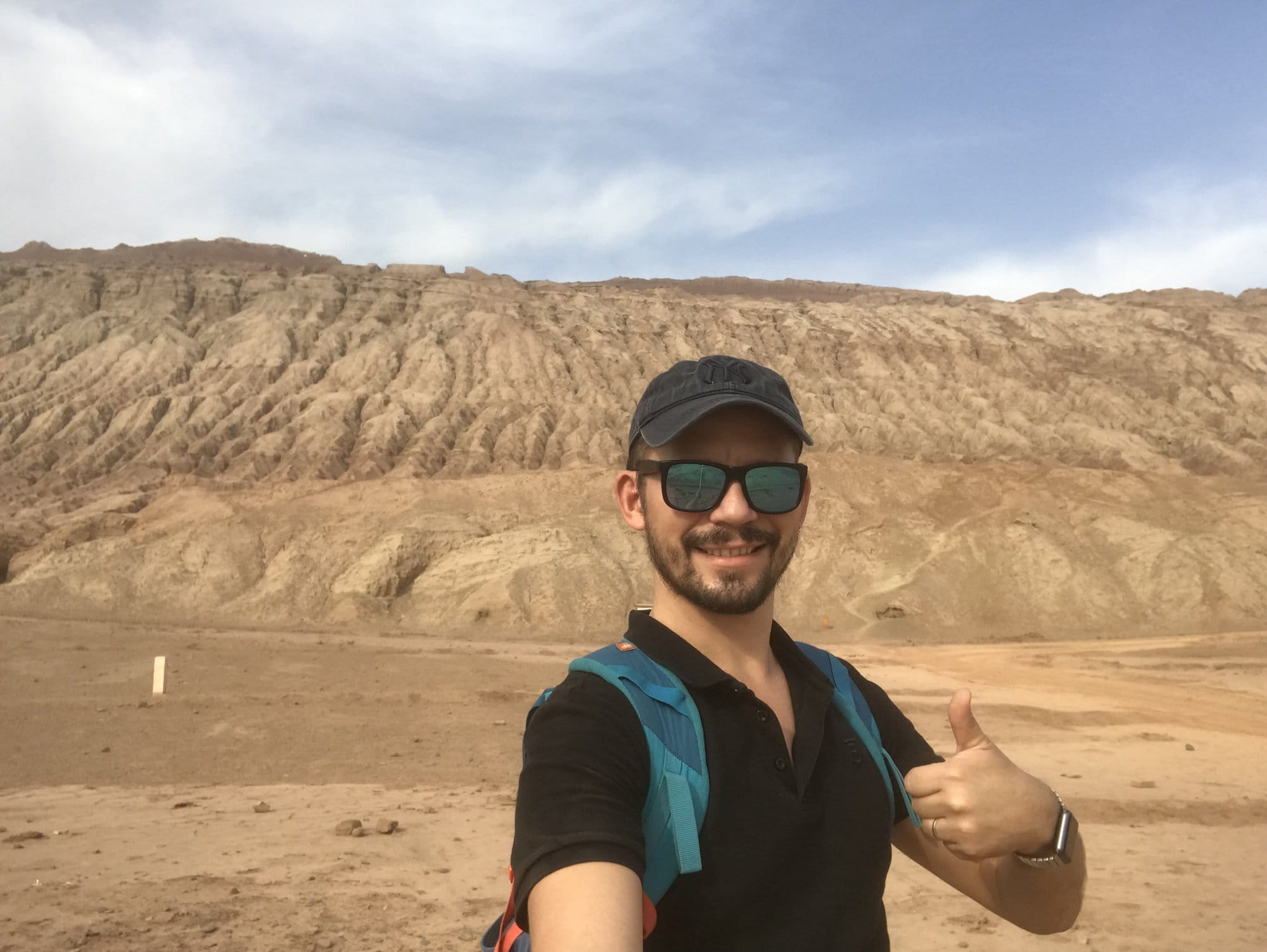 Thumbs up in front of a striped earth cliff and the desert plateau beyond.