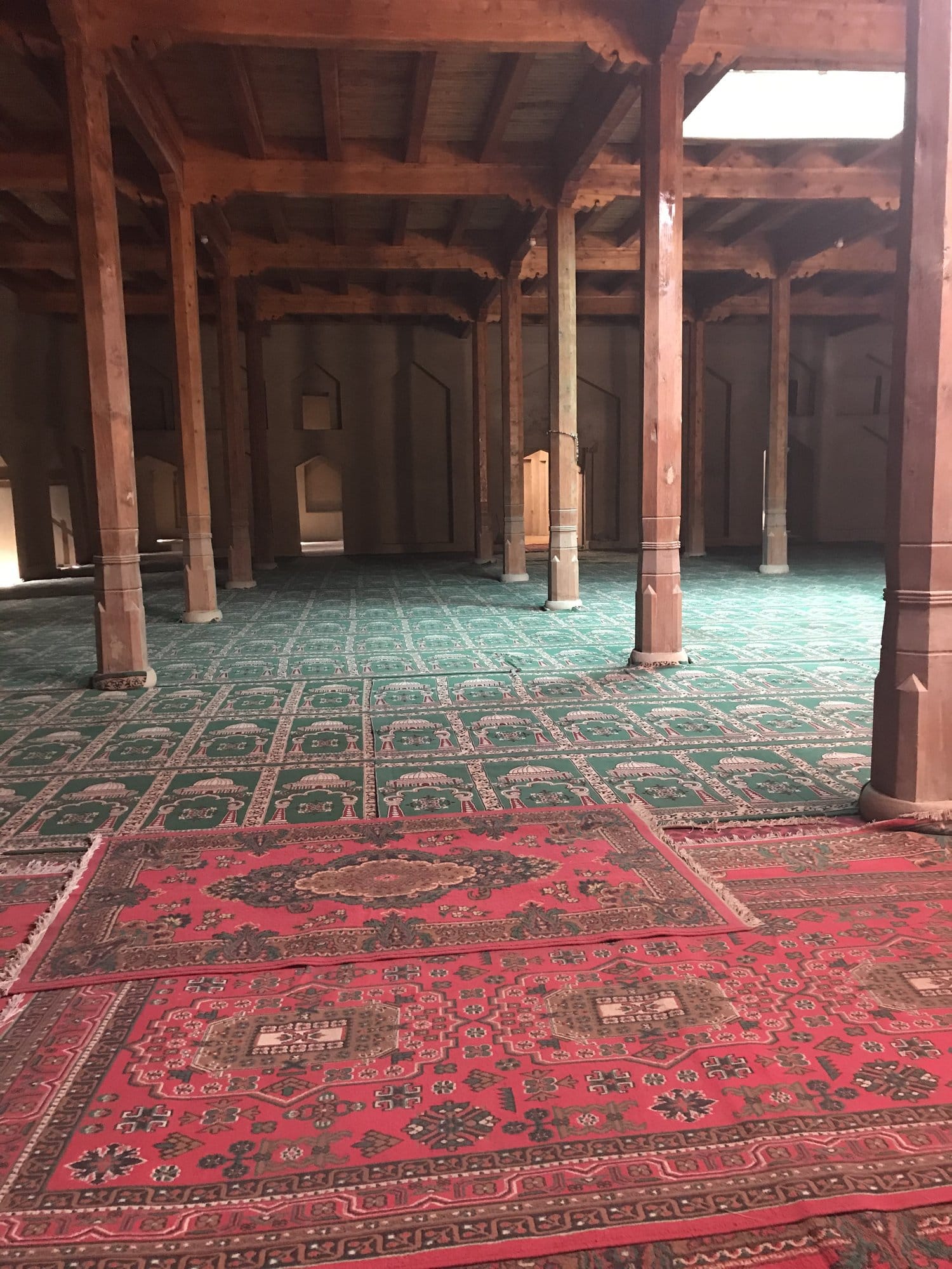 Inside a wood-columned prayer hall, red carpets on the floor and green matting patterned with mihrabs.