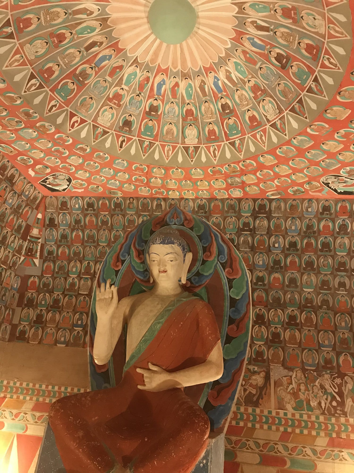 A painted Buddha seated beneath a dome covered in hundreds of smaller Buddhas, the frescoes visibly damaged.