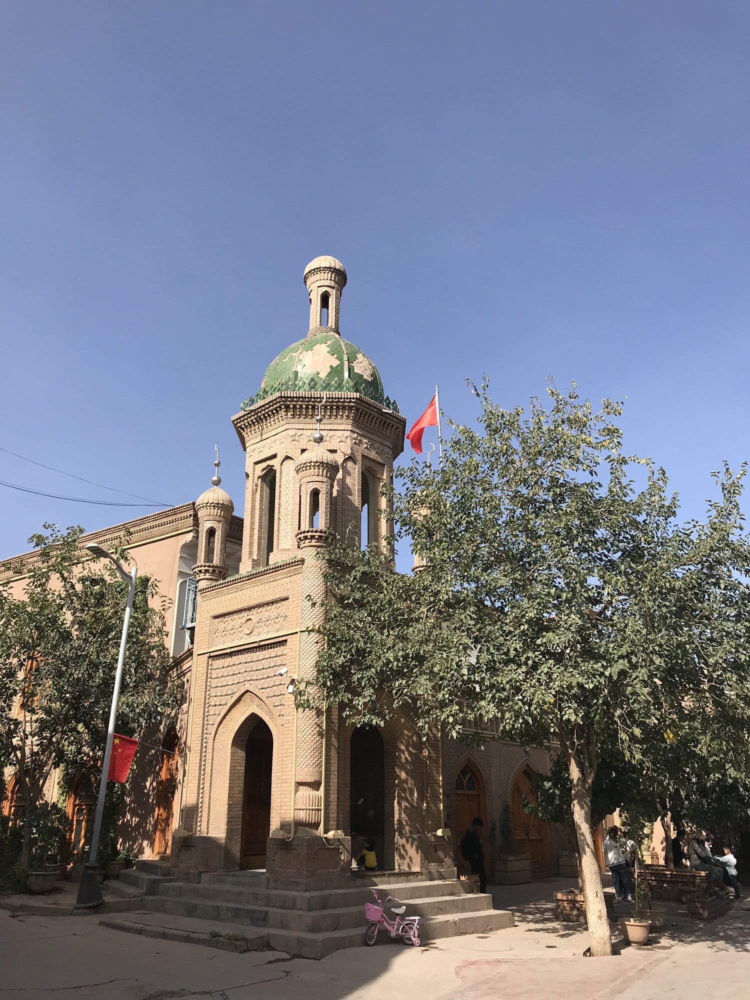 A neighbourhood mosque in brick with a green dome and minaret, a child's pink bicycle left at the foot of the steps.