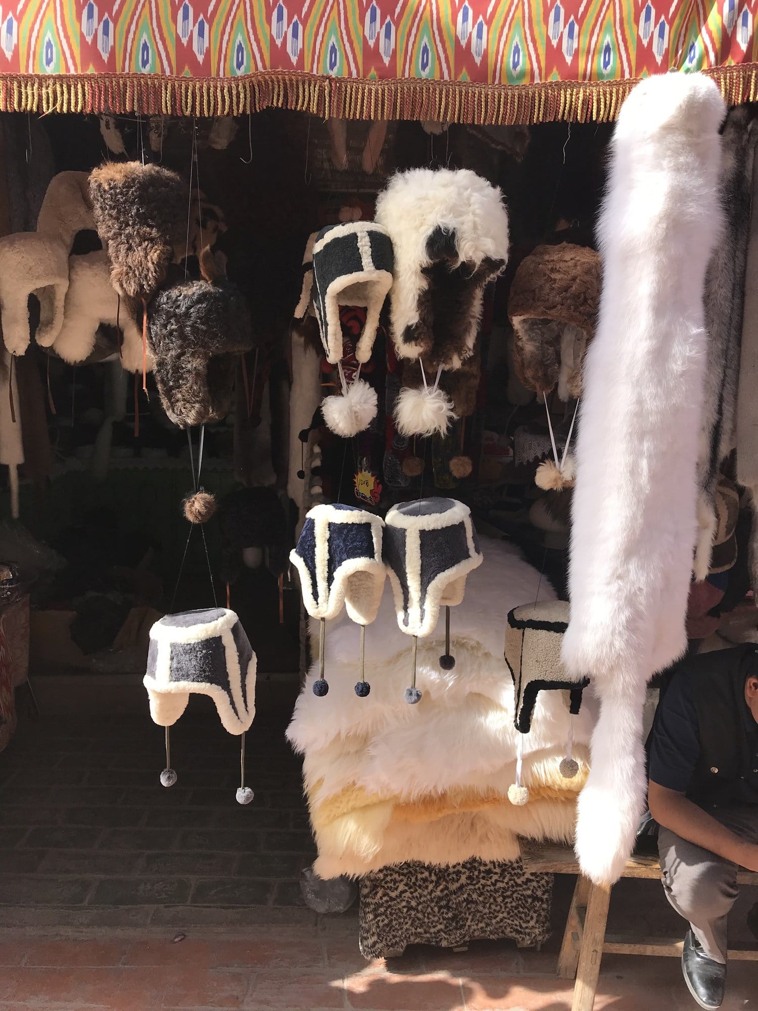 Fur hats and caps hung under an ikat-patterned awning, white skins stacked below.