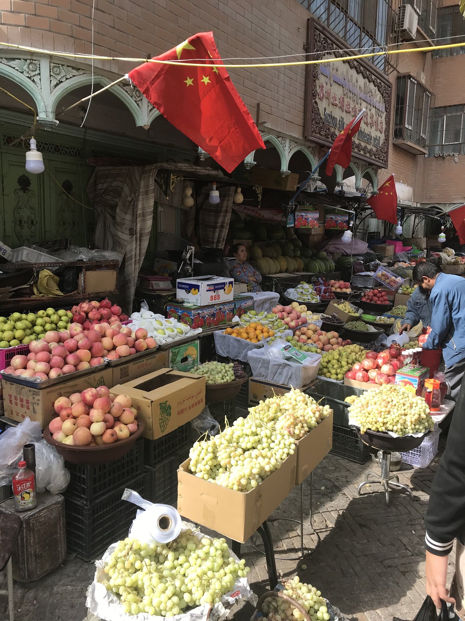 The covered fruit market, crates of apples, white grapes, pomegranates and melons under bare bulbs.