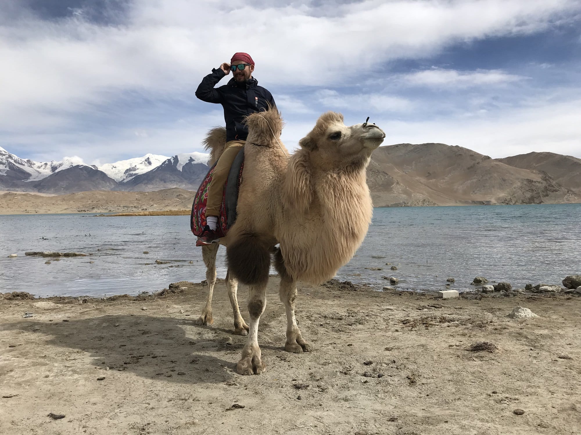 Riding a Bactrian camel at the edge of Karakul, the snow mountains behind.