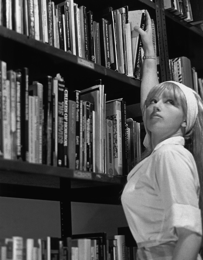 A blonde woman wearing a white shirt reaches above her head toward a bookshelf. 